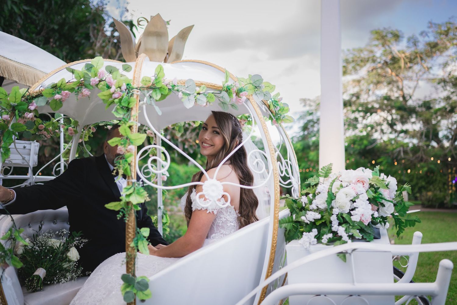 Smiling bride sitting in a decorated white Cinderella carriage at outdoor wedding.