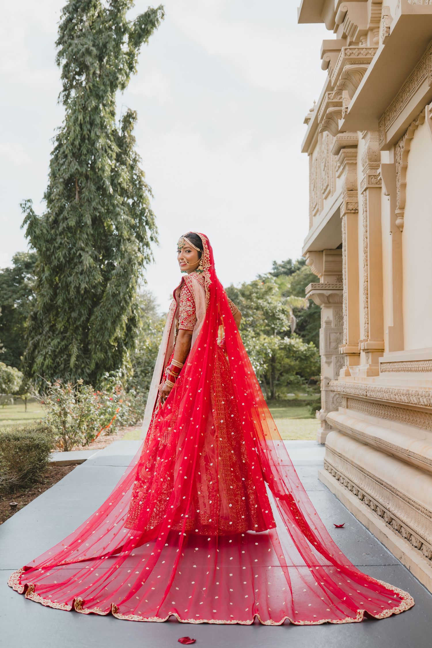 Indian bride in stunning red lehenga with embroidered veil poses gracefully outside ornate temple.