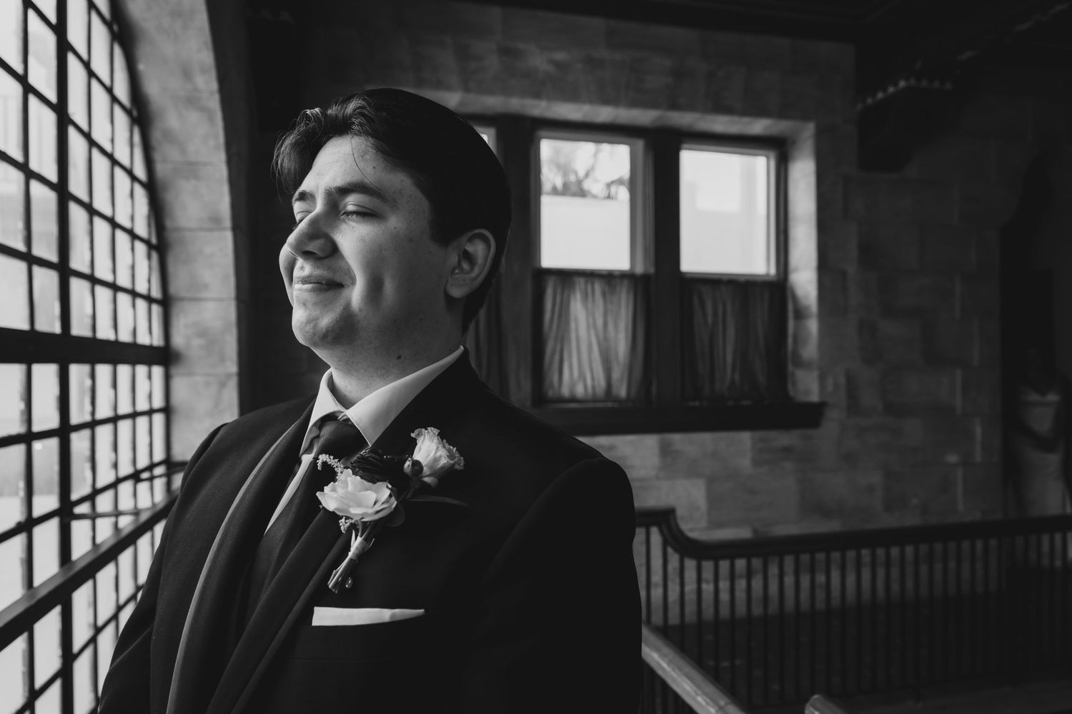 Black and white portrait of a groom in a suit with boutonniere, gazing thoughtfully near a window.
