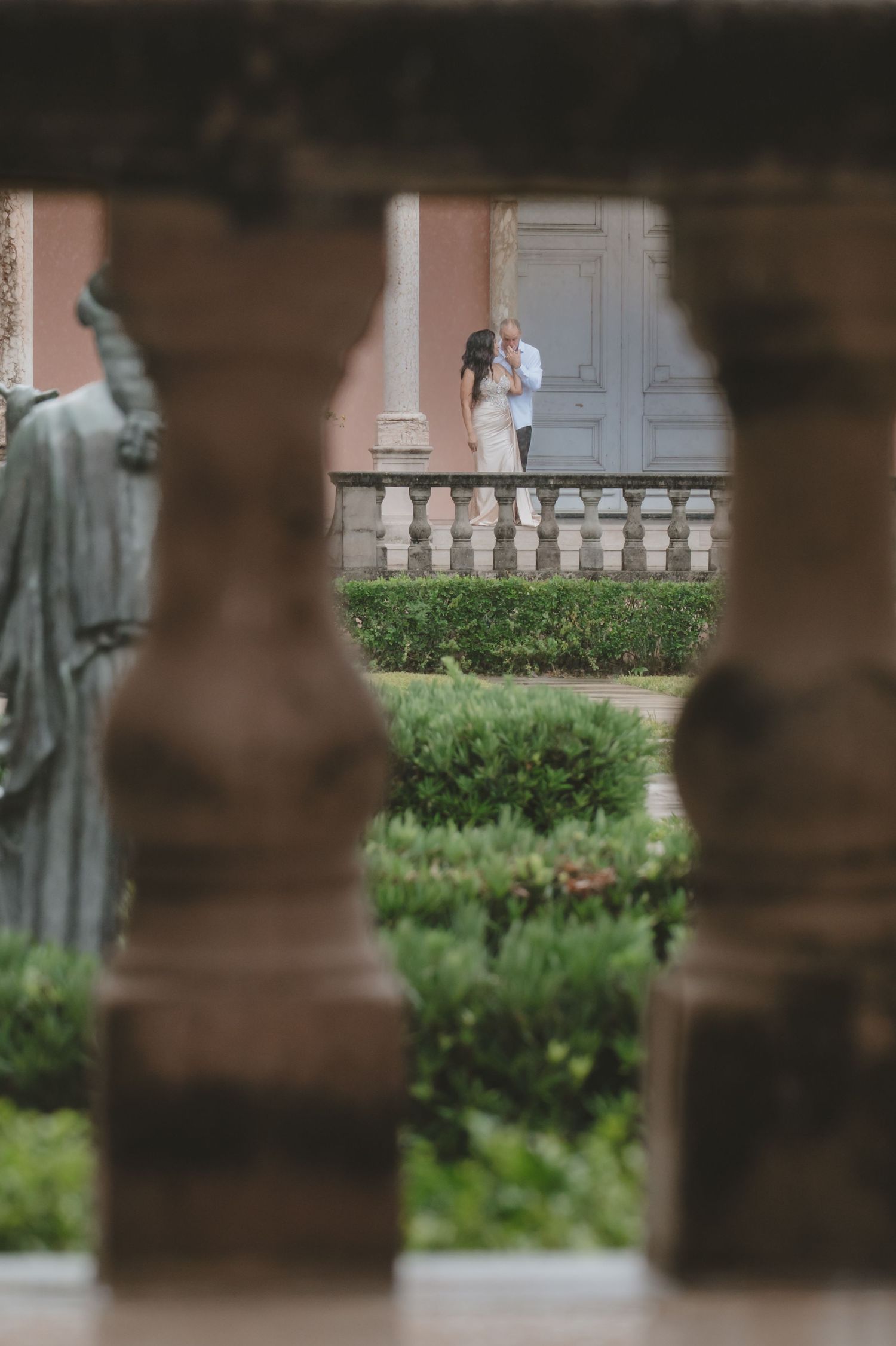 Couple embracing on a balcony framed by stone balusters and lush garden hedges at an elegant villa.