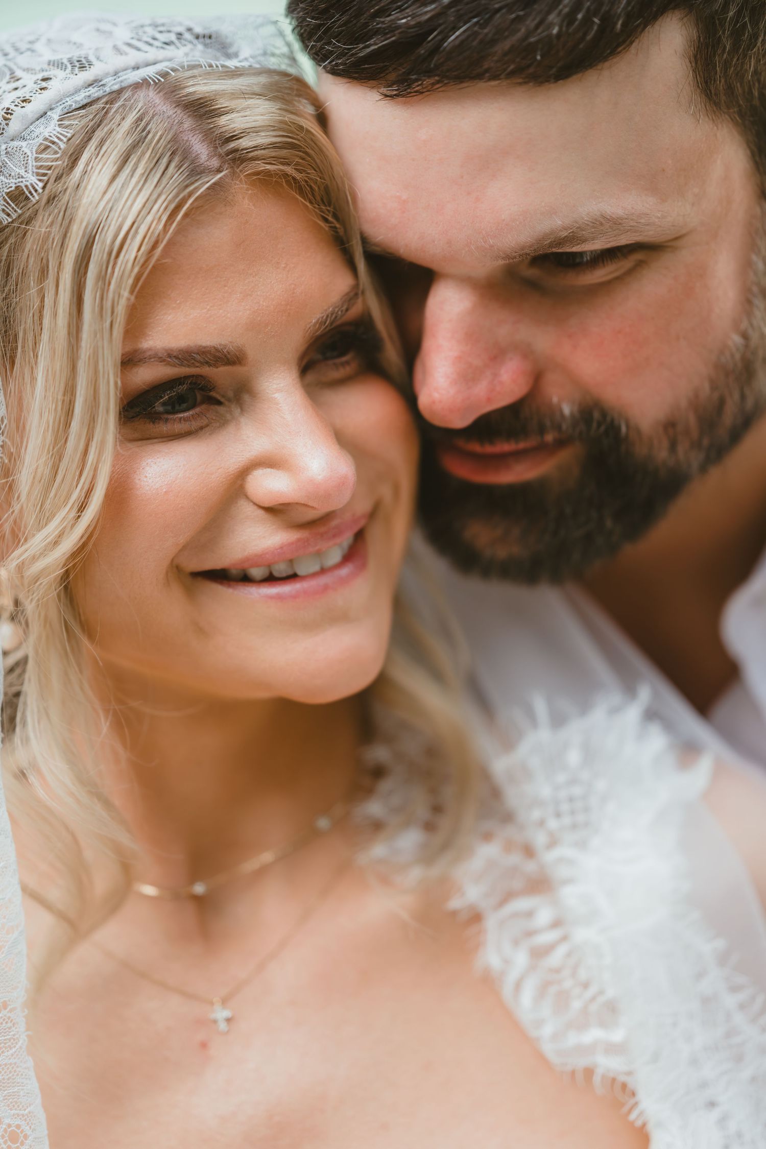 Close-up portrait of a smiling bride with a lace veil as her groom tenderly nuzzles her cheek.