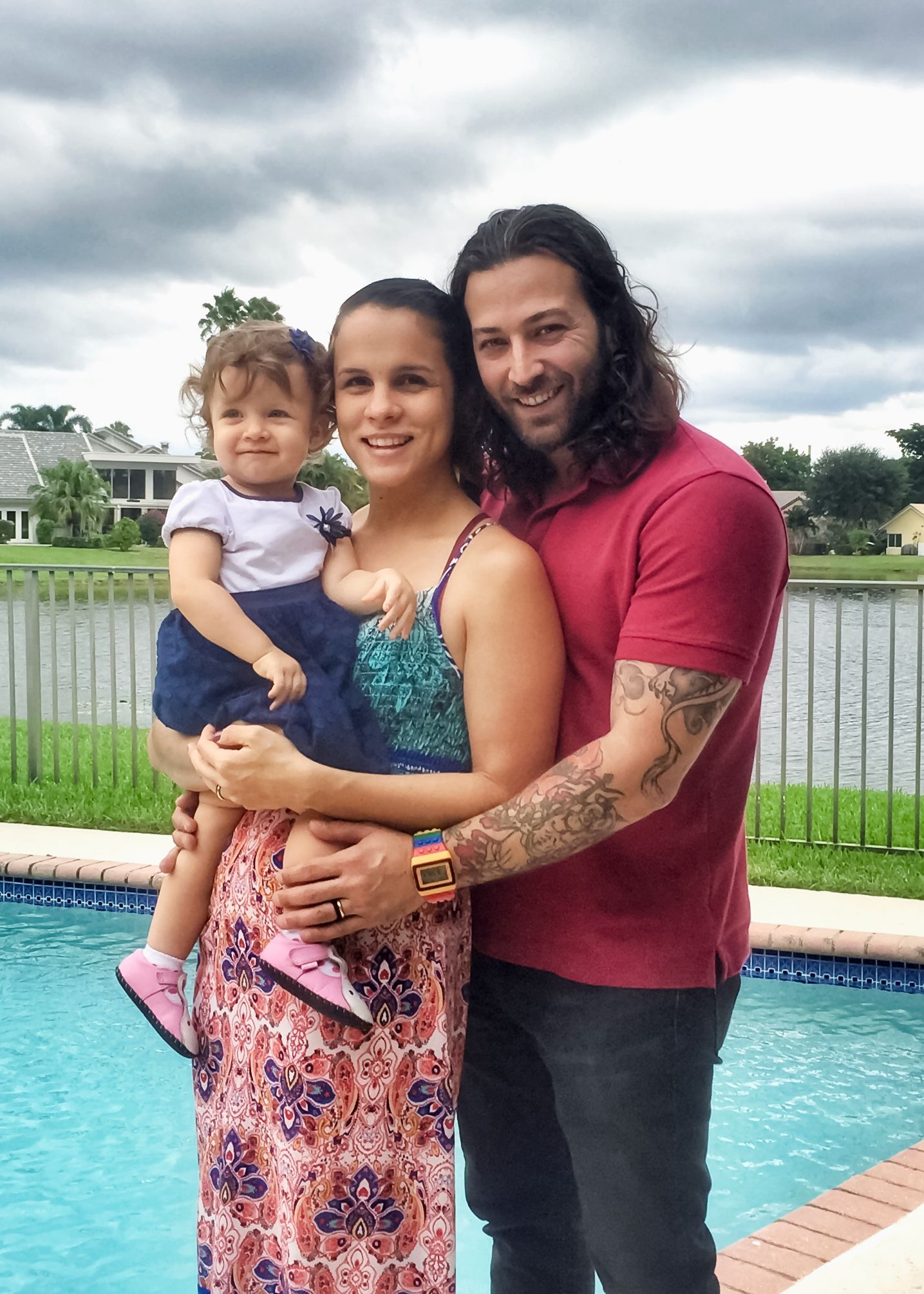 Happy family of three posing by pool, man with tattoos and long hair, woman holding toddler.