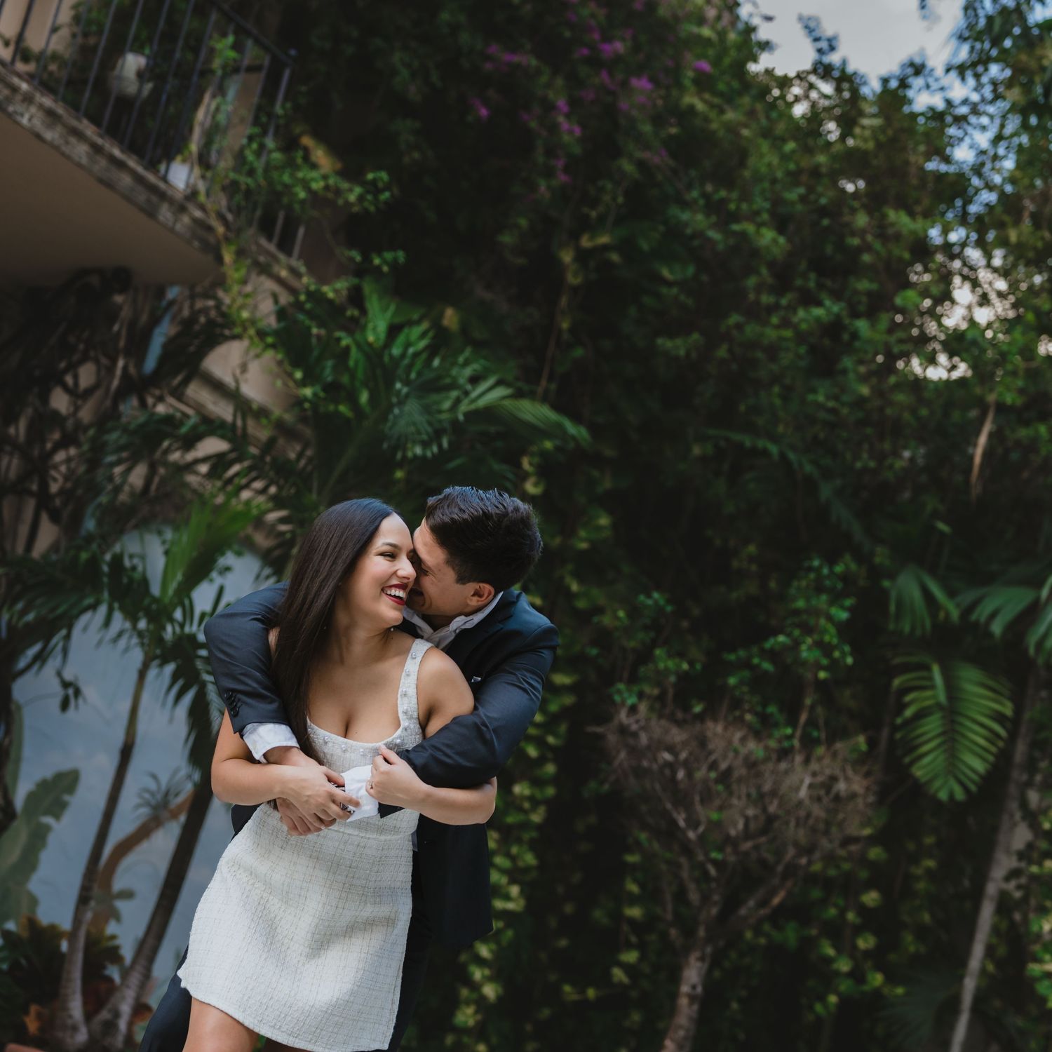 Man embracing woman from behind as she smiles joyfully surrounded by tropical greenery.