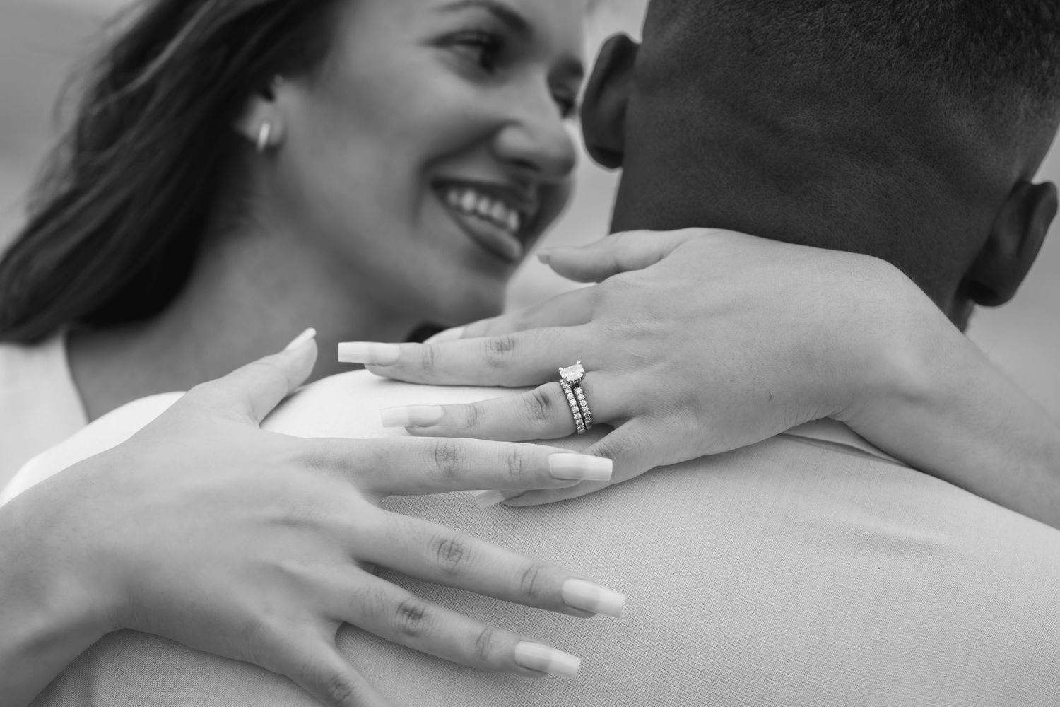 Close-up black and white photo of smiling bride showing diamond engagement ring while hugging groom.