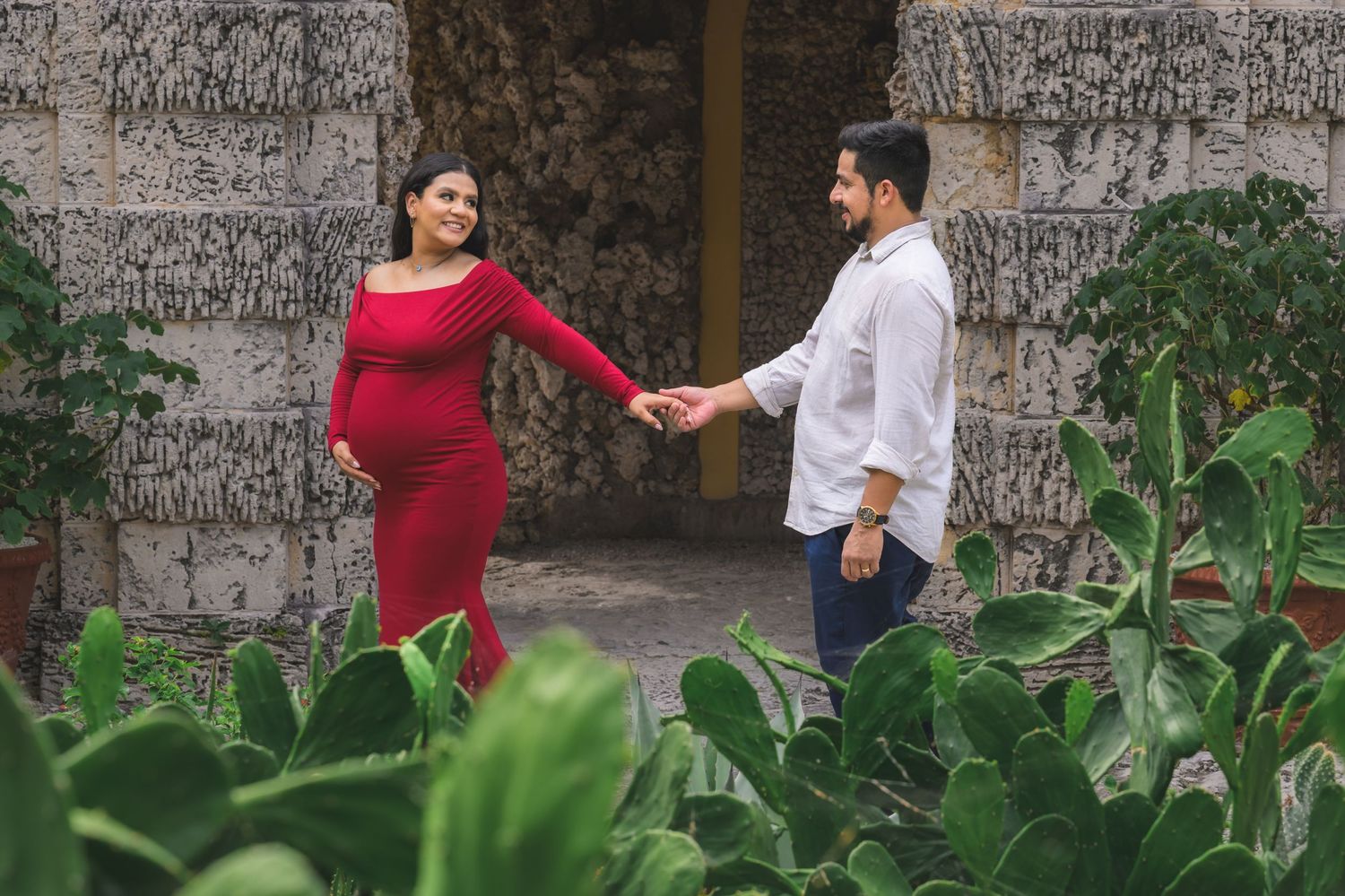 Pregnant woman in red dress holds hands with man in white shirt outdoors among cacti and stone columns.