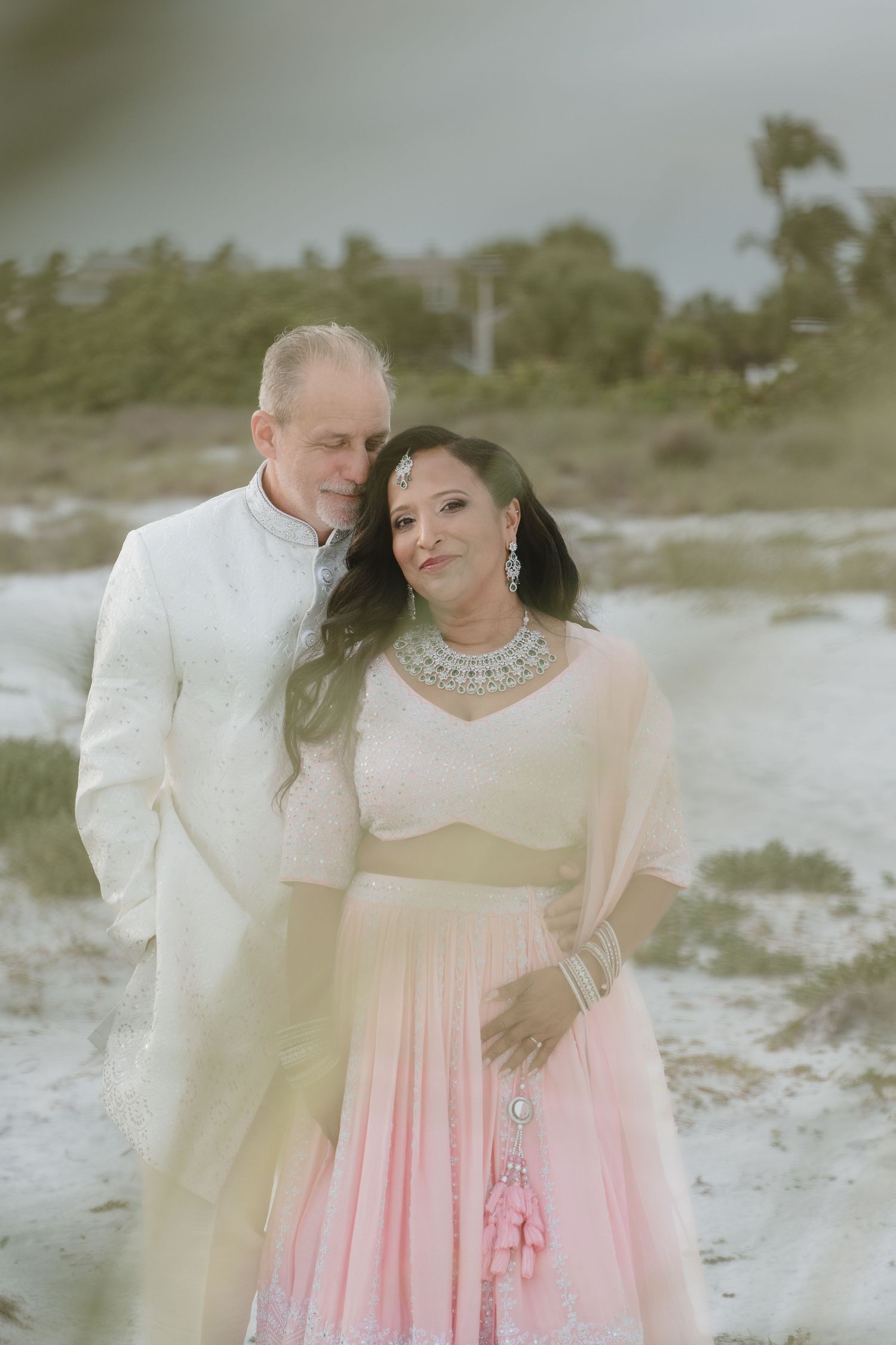Couple in Indian wedding attire on sandy beach, man in white sherwani, woman in pink lehenga with jewelry.