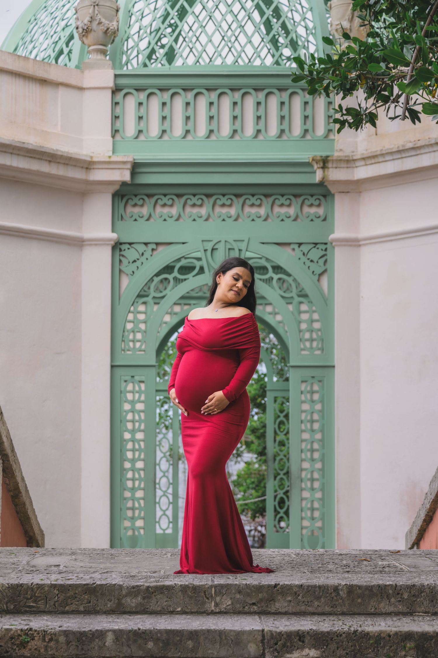Pregnant woman in red off-shoulder gown posing before ornate green architectural backdrop.