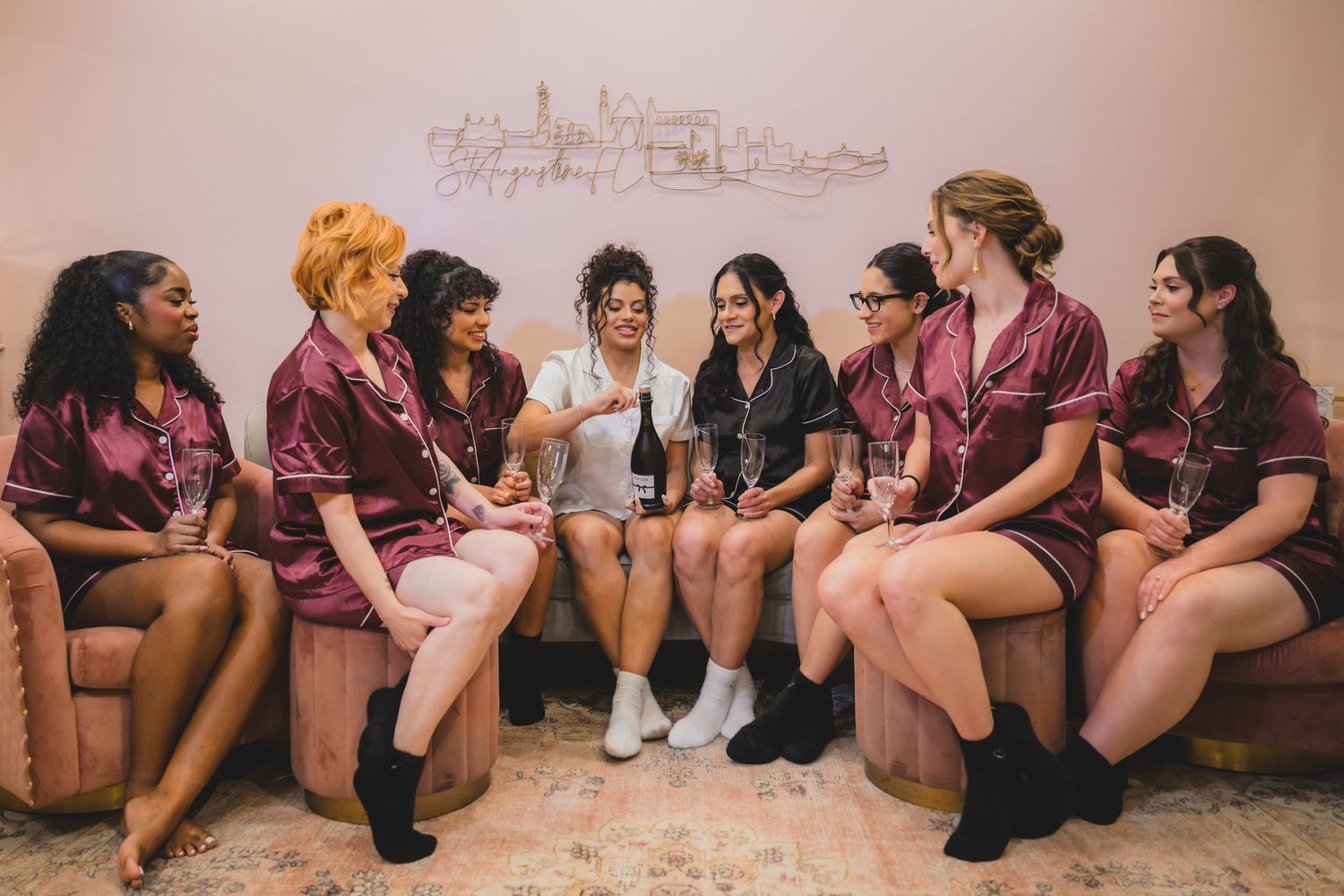 A group of bridesmaids in matching burgundy satin robes sitting together on a pink sofa, laughing and celebrating.