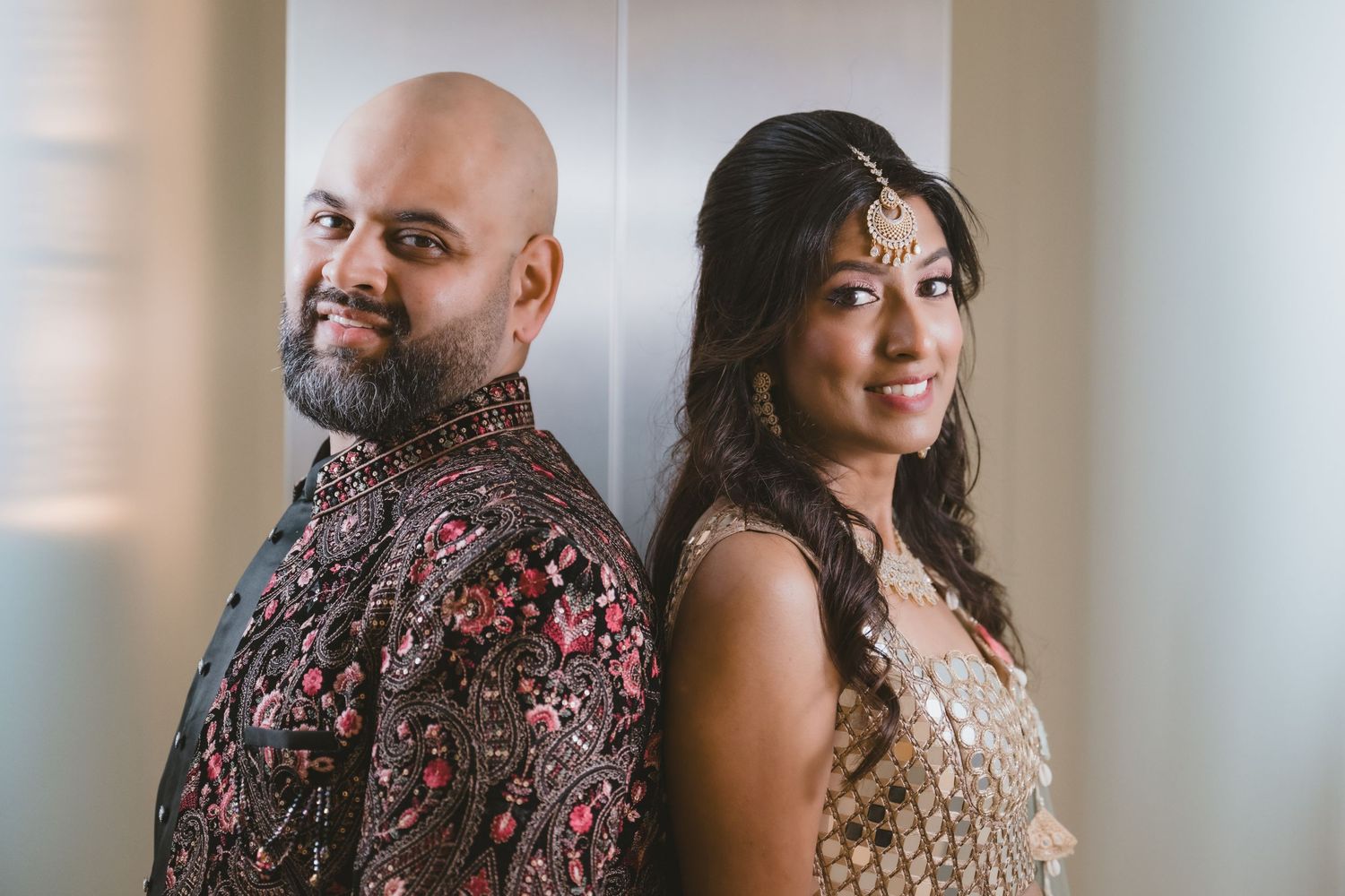 Indian couple in ornate traditional attire standing back to back smiling in elegant portrait.