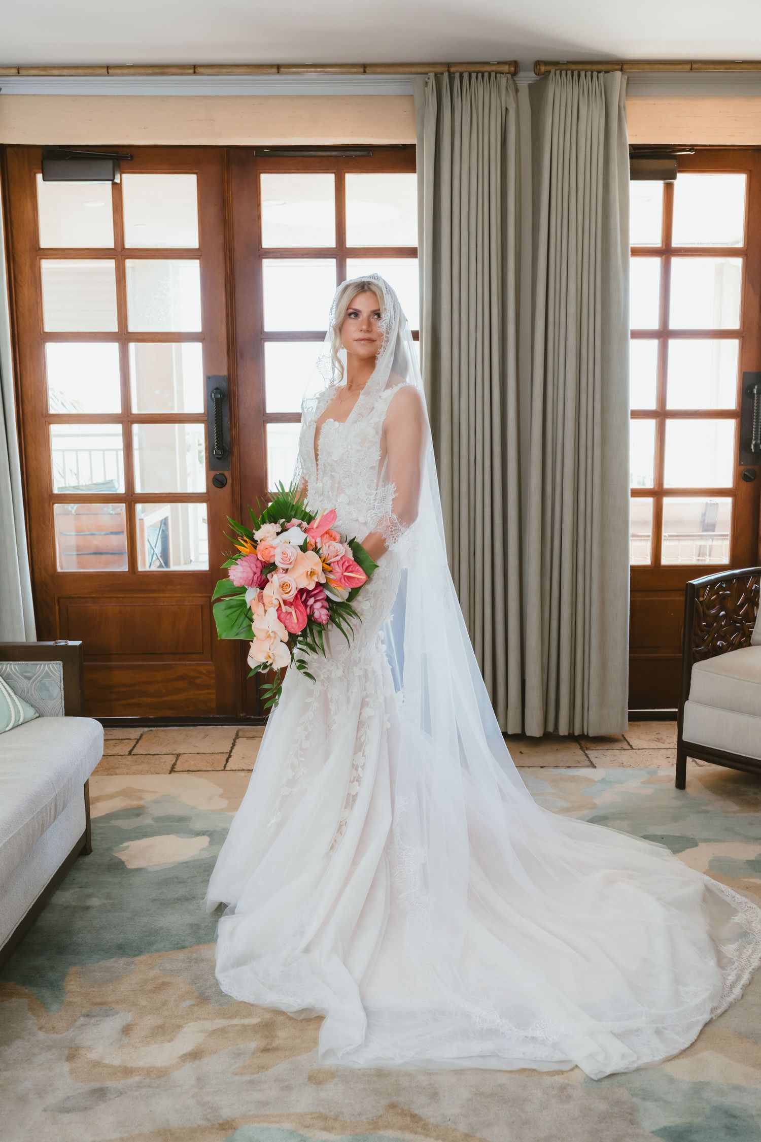 Blonde bride in lace wedding gown with veil holding colorful pink tropical bouquet indoors.