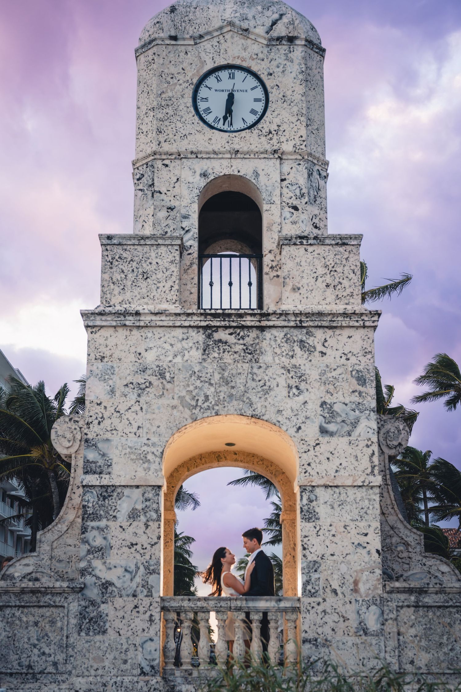 Wedding couple posing under historic clock tower arch with dramatic purple sunset sky and palm trees.