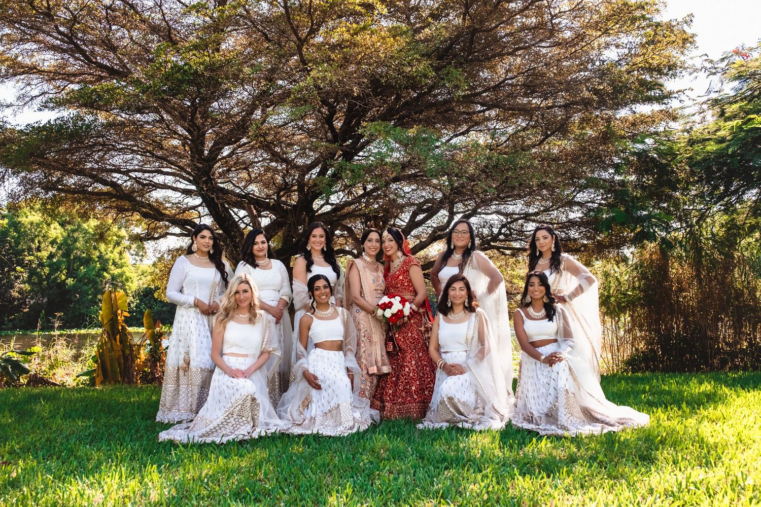 Bride in red lehenga posing with bridesmaids in white dresses under a large tree on a sunny day.