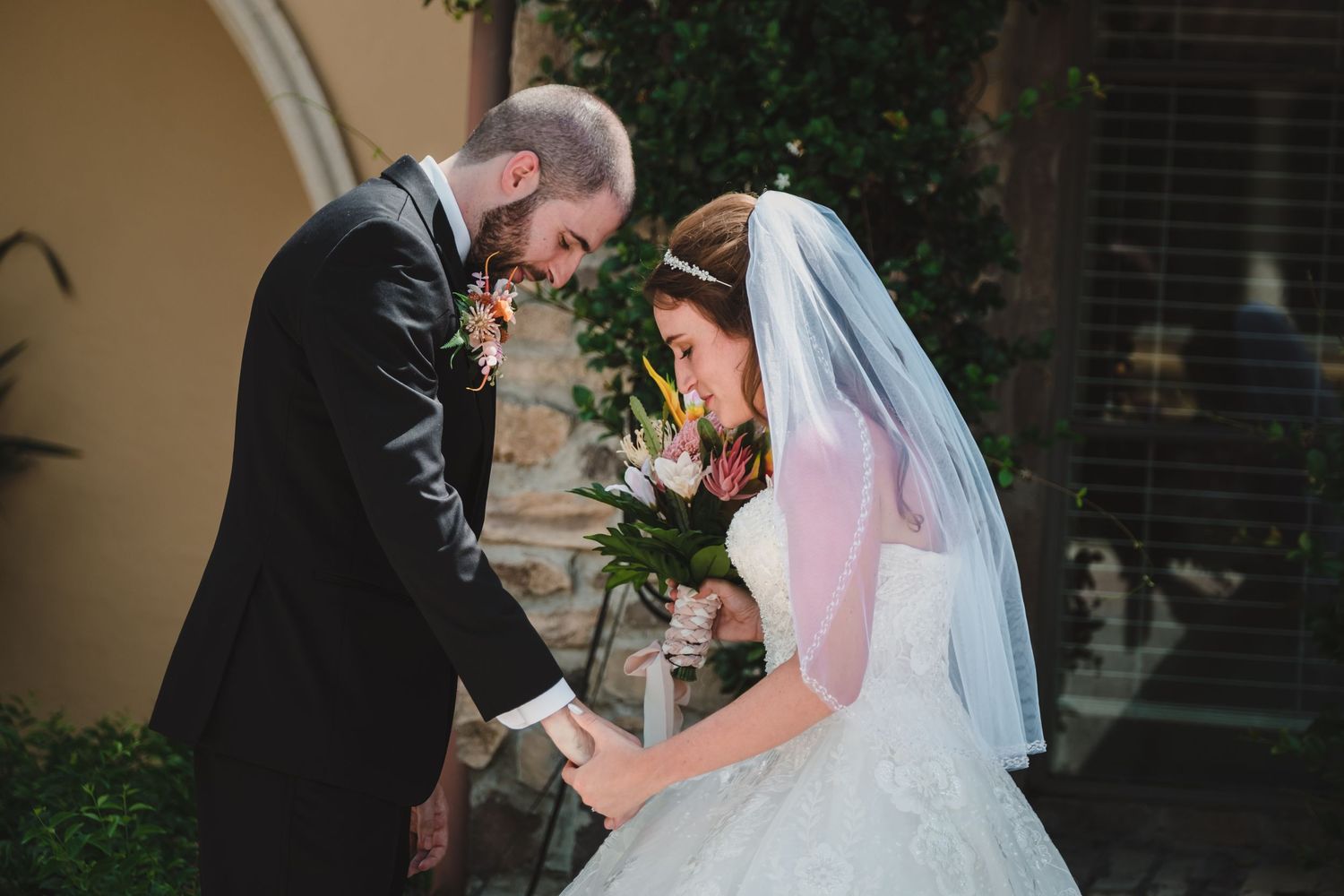 Bride and groom share a tender bowed-head moment while holding hands and colorful bouquet outdoors.