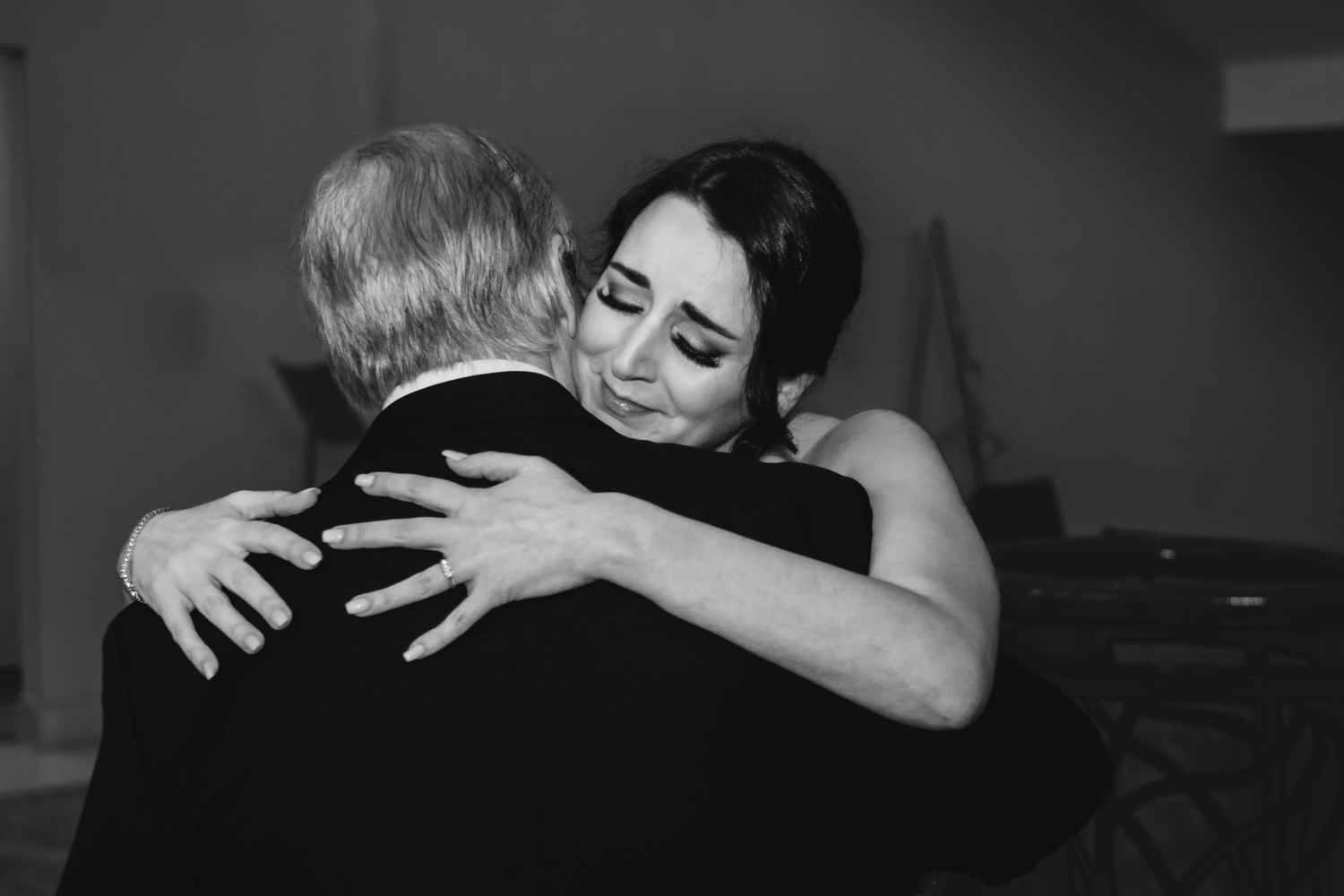 Emotional bride shares tender hug with older man in black tuxedo at wedding reception.
