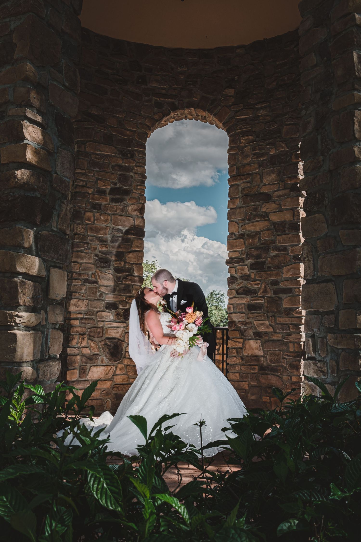 Wedding couple shares a romantic kiss inside a rustic stone tower with an arched window.
