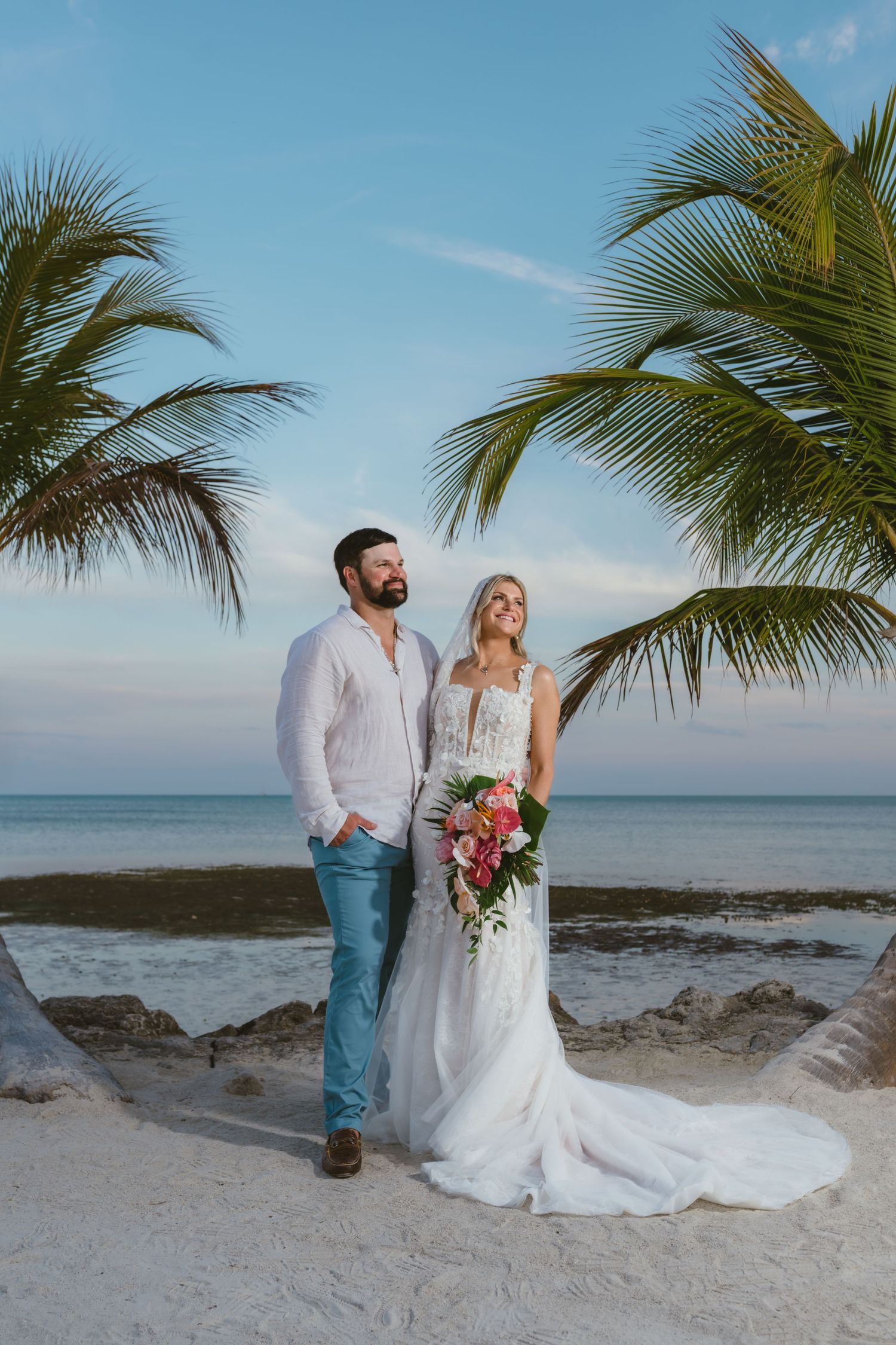 Beach wedding couple stands under palm trees at sunset with tropical bridal bouquet.