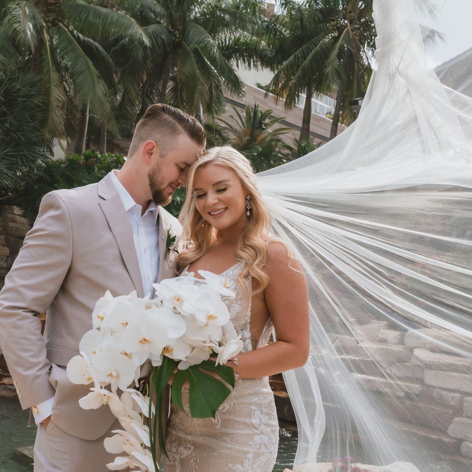 Bride and groom share romantic moment with flowing veil and white orchid bouquet surrounded by tropical palms.
