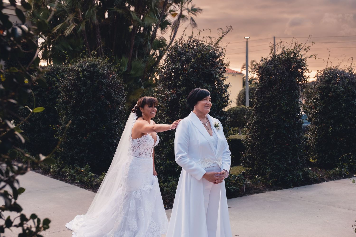 Bride in white lace gown with veil walks alongside woman in white suit at sunset.