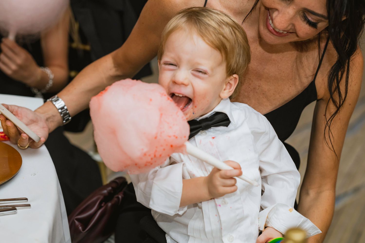 Adorable toddler boy in bow tie joyfully eating pink cotton candy at a wedding reception.