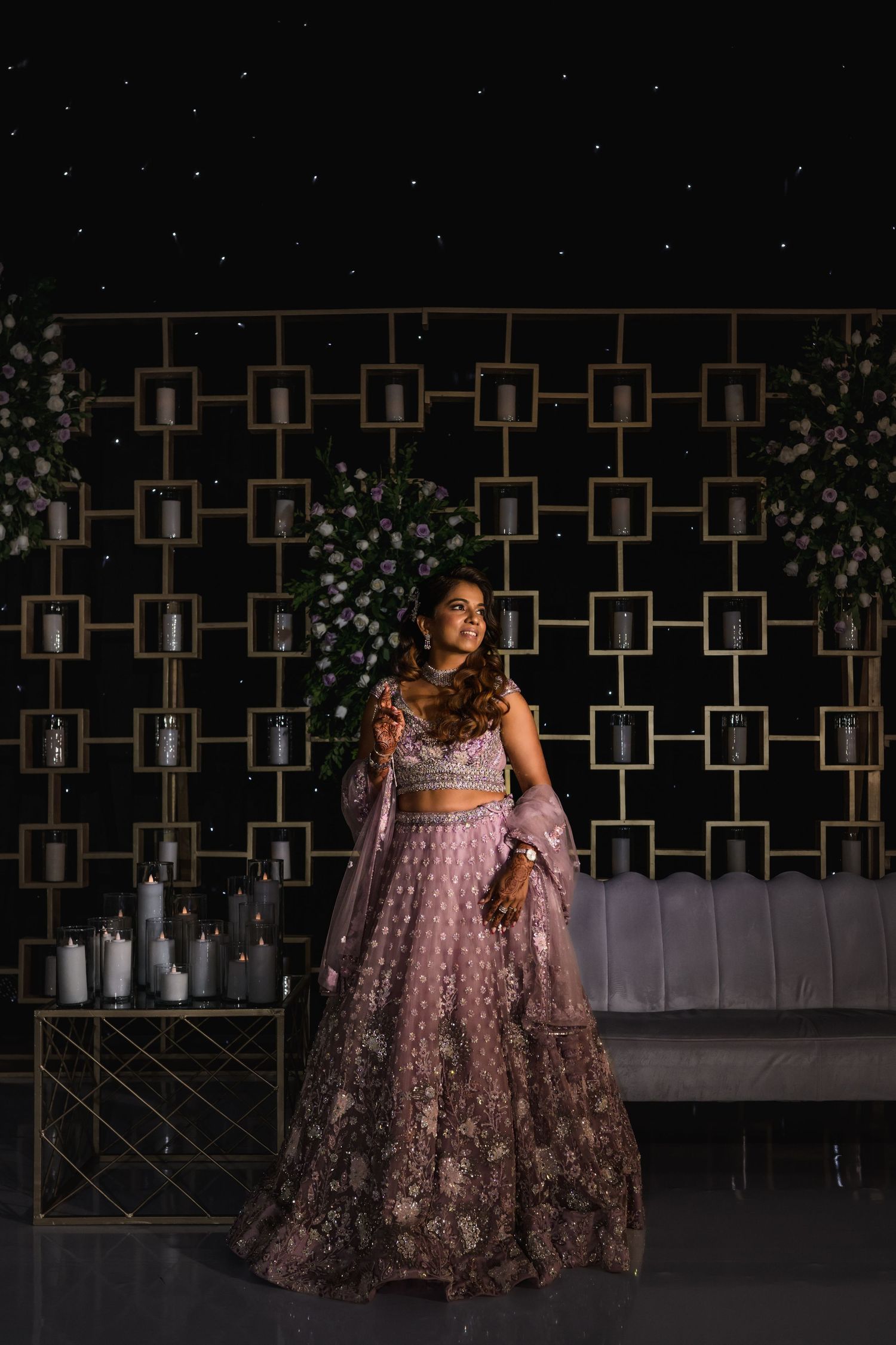 Bride in dusty rose lehenga poses elegantly against geometric candle backdrop at wedding reception.