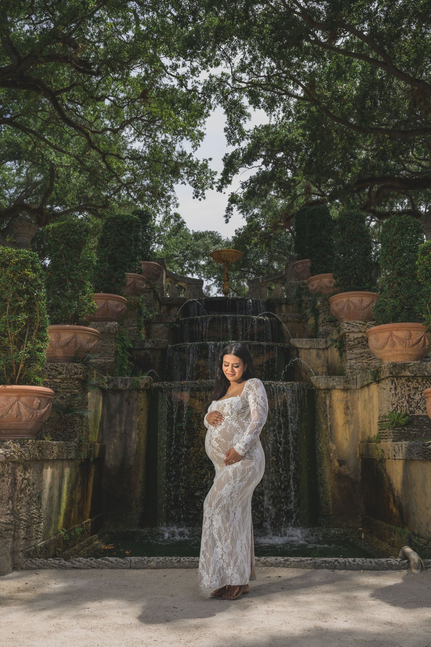 Pregnant woman in white lace gown poses before a garden waterfall surrounded by lush greenery and terracotta urns.