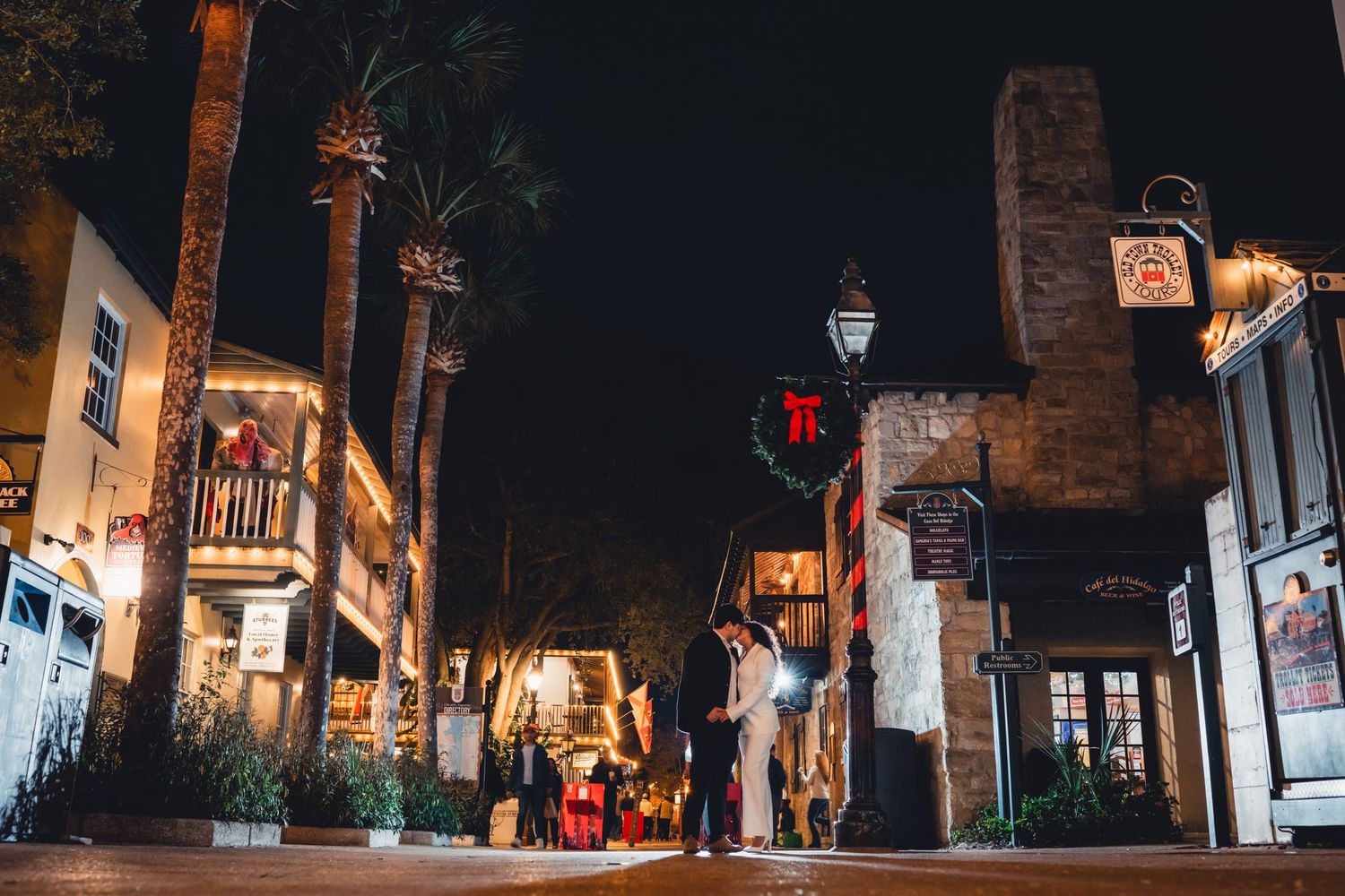 Historic St. Augustine street at night with Christmas wreaths, palm trees, and festive lights illuminating stone buildings.