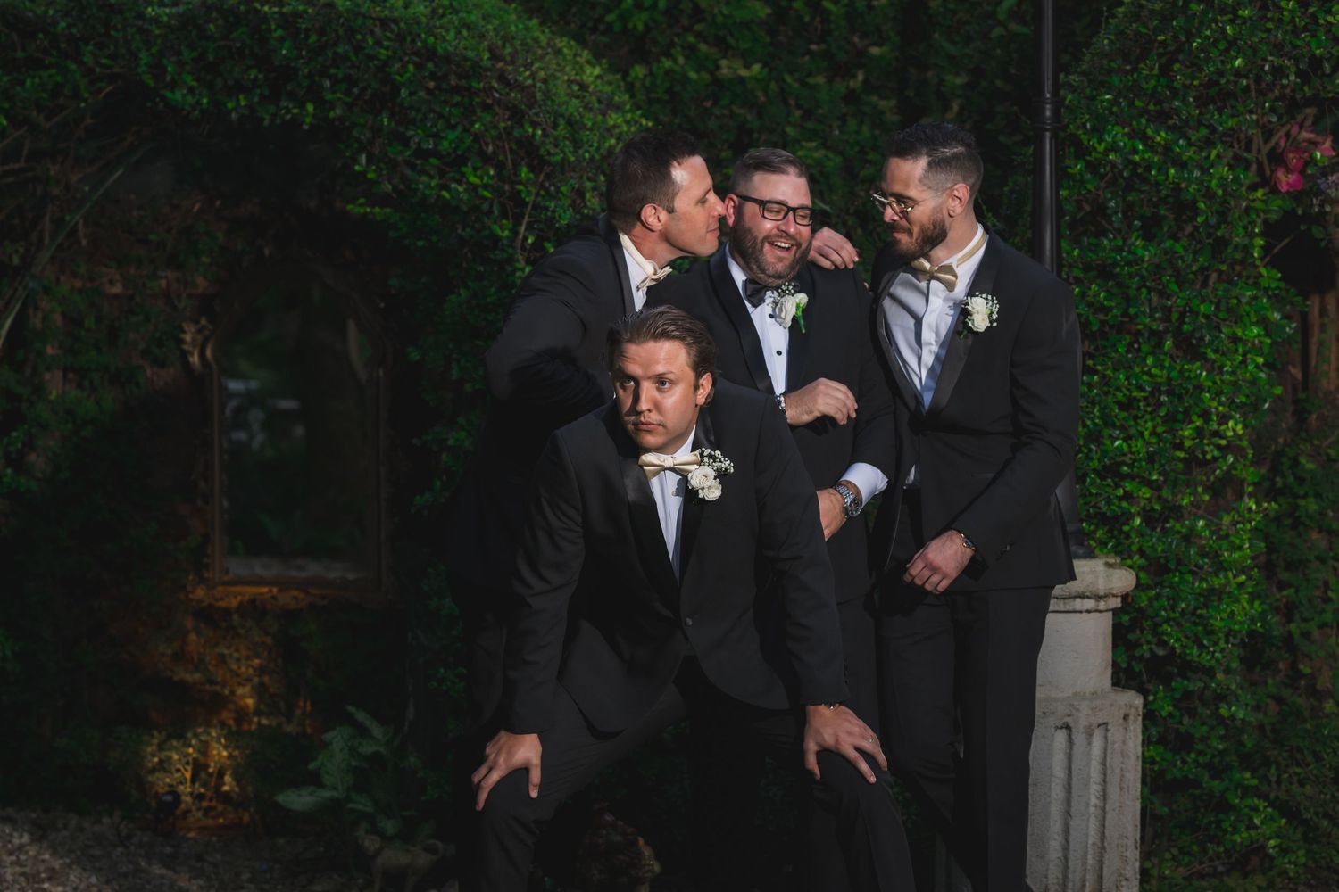 Groomsmen in black suits laughing and posing playfully against a lush green ivy wall outdoors.