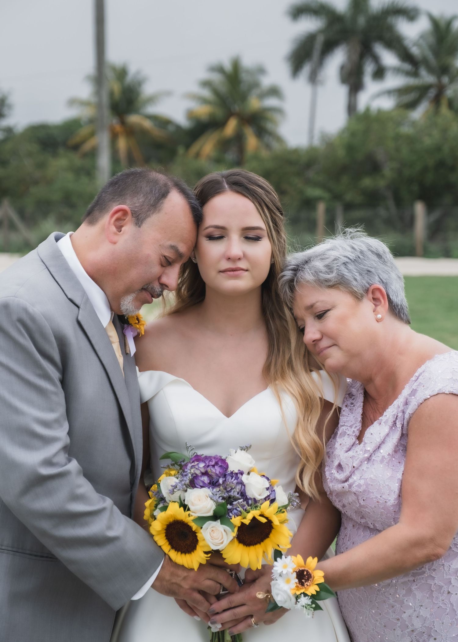 Bride with parents bowing heads in prayer holding sunflower and white rose bouquet outdoors.