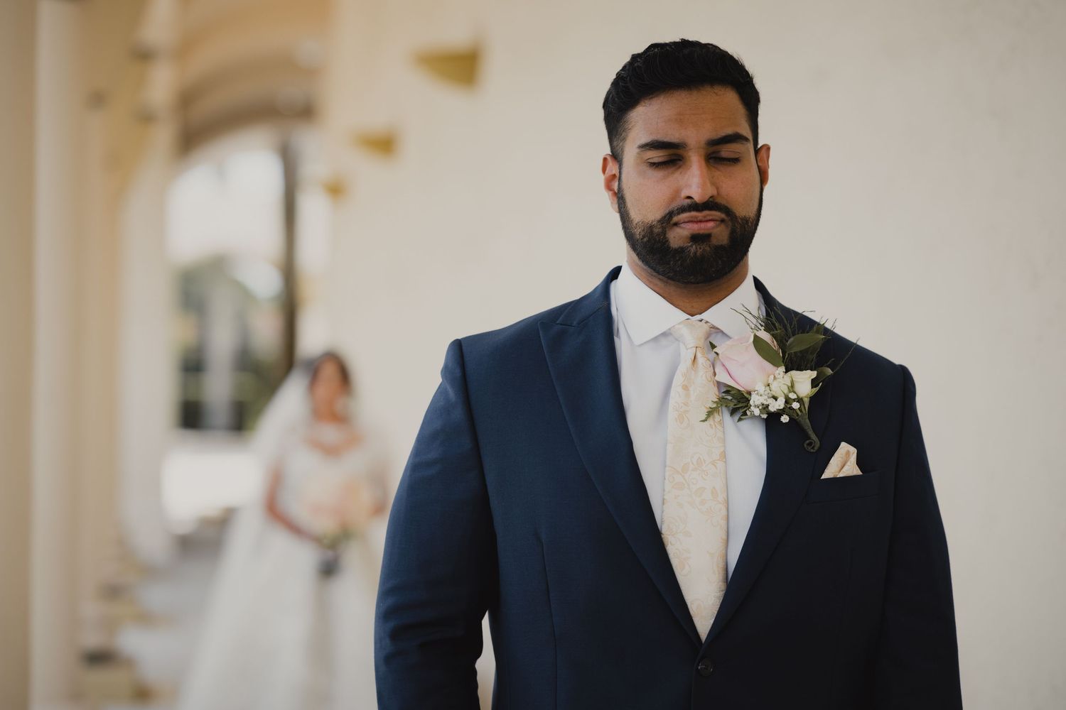 Groom in navy suit with floral boutonniere eyes closed before first look wedding ceremony.