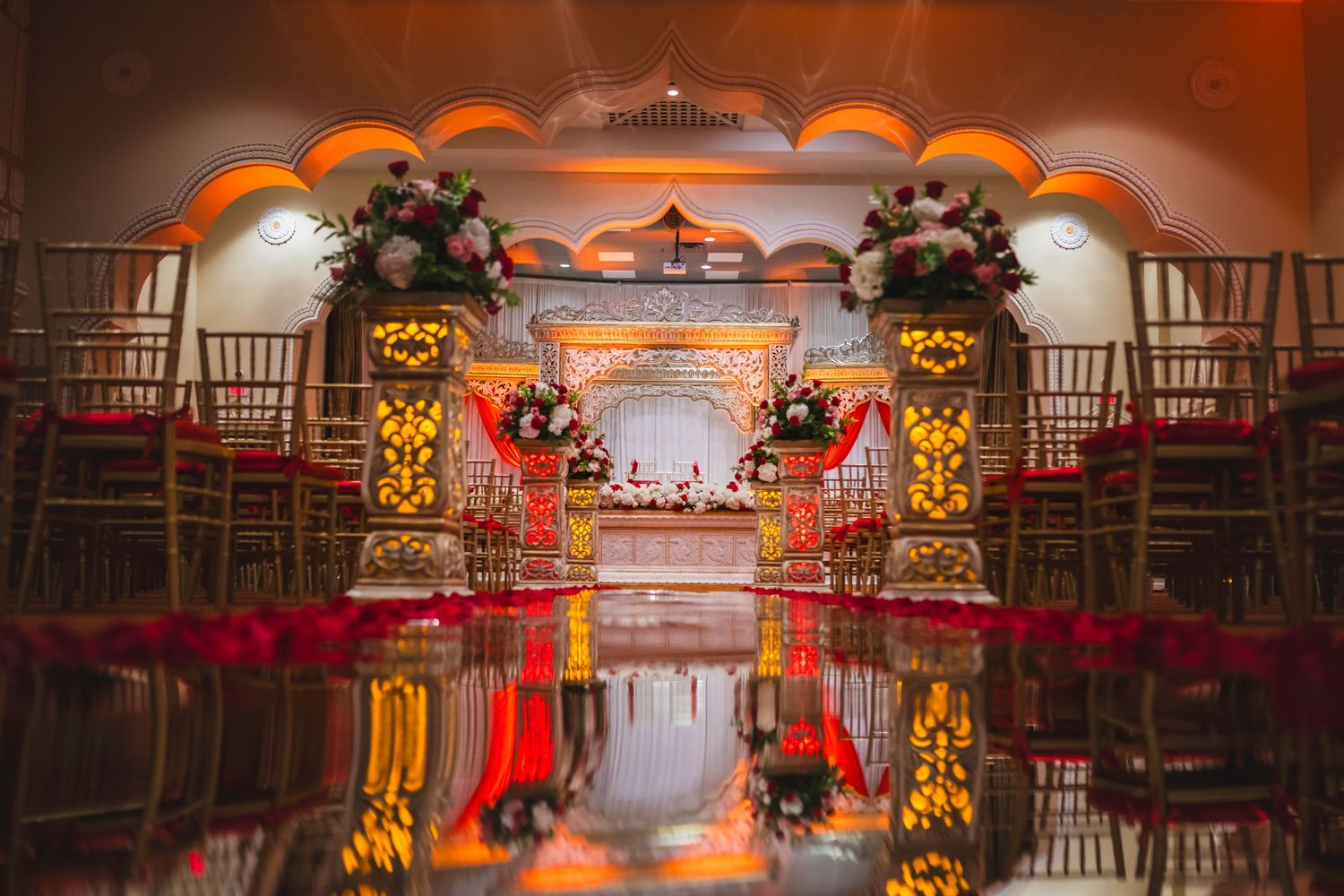 Elegant Indian wedding venue with reflective floor, golden arches, floral arrangements, and red carpet aisle.