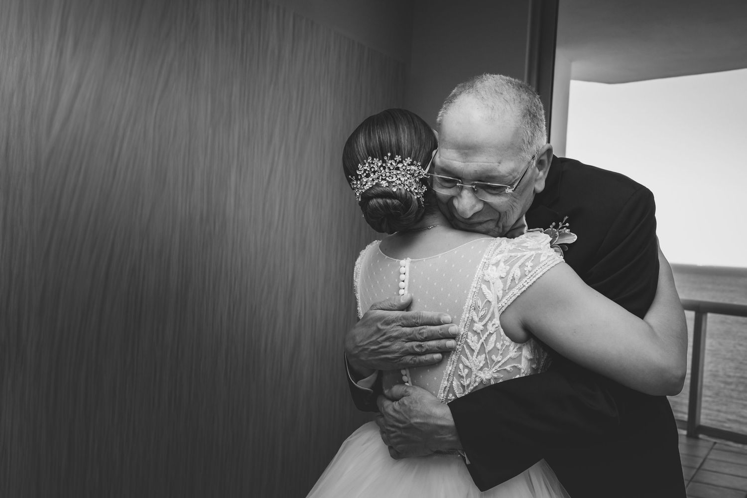 Black and white photo of a bride in a lace gown embracing an older man, sharing a tender moment on her wedding day.