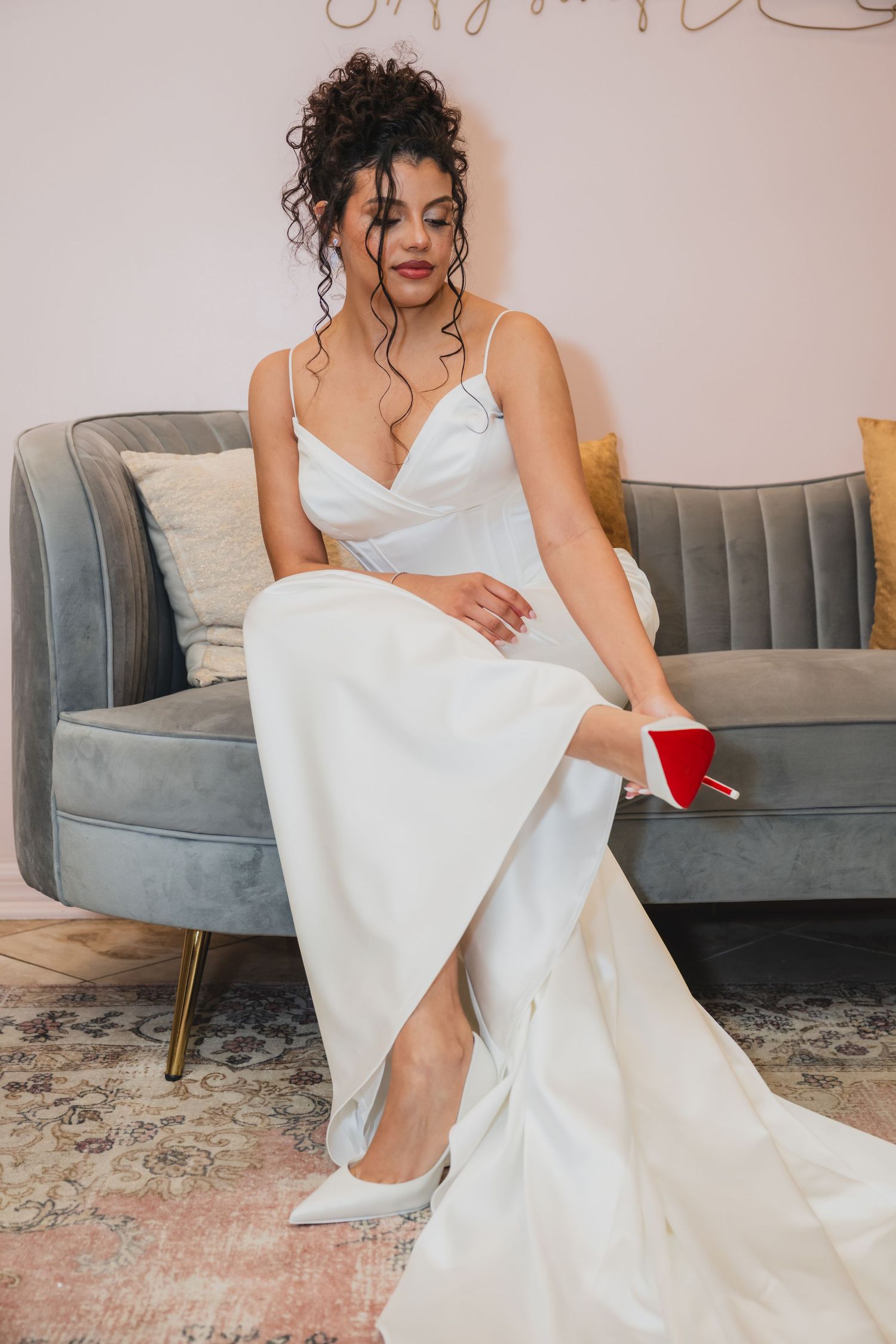 Bride in elegant white satin gown sitting on grey velvet sofa adjusting red-soled Louboutin heels.