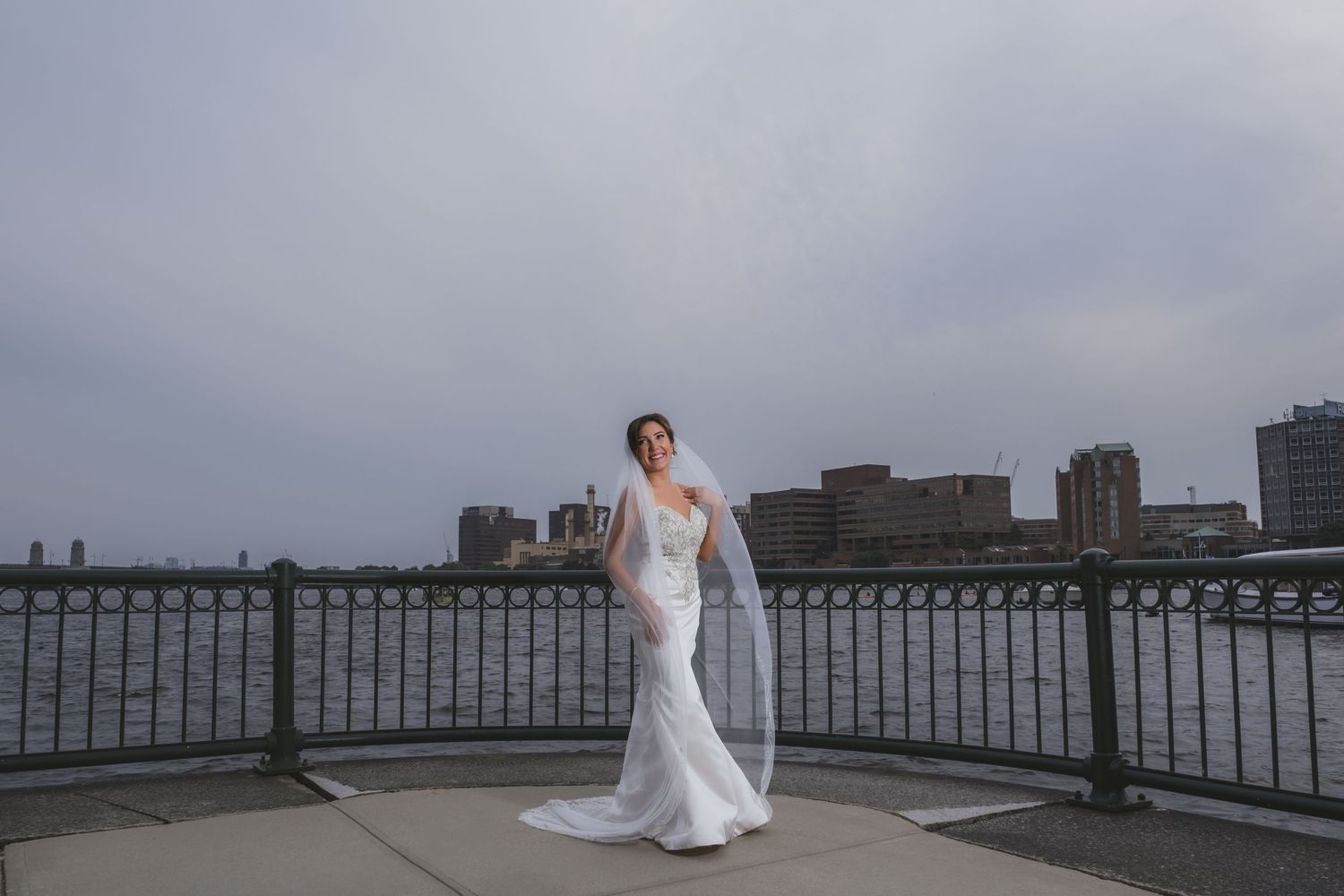 Elegant bride in fitted white gown with veil posing by waterfront railing against city skyline.