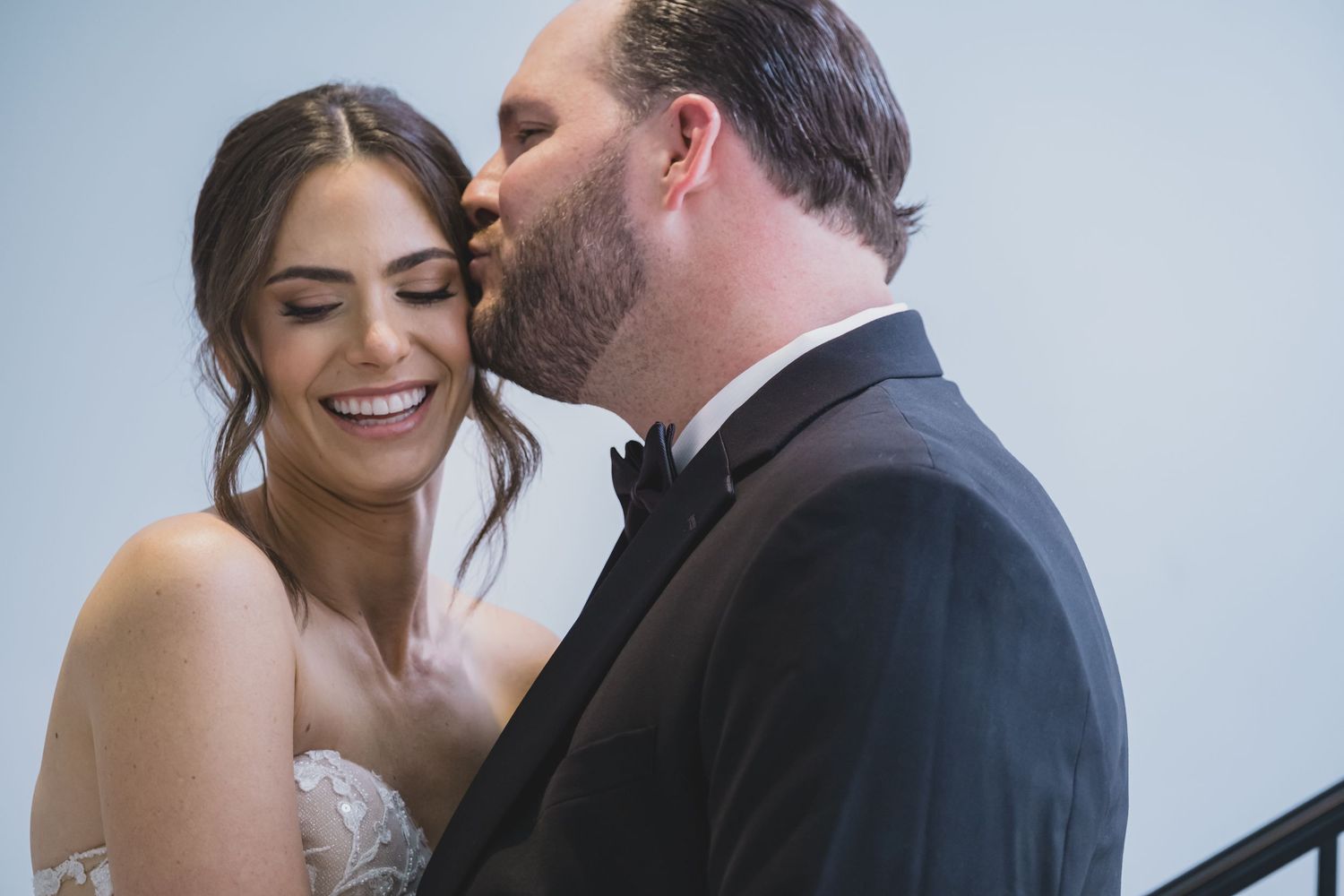 Groom in black tuxedo kisses smiling bride on the cheek in romantic indoor wedding portrait.