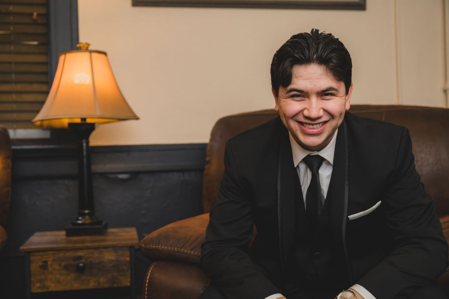 Smiling young man in black suit and tie seated on leather chair beside lamp in elegant room.