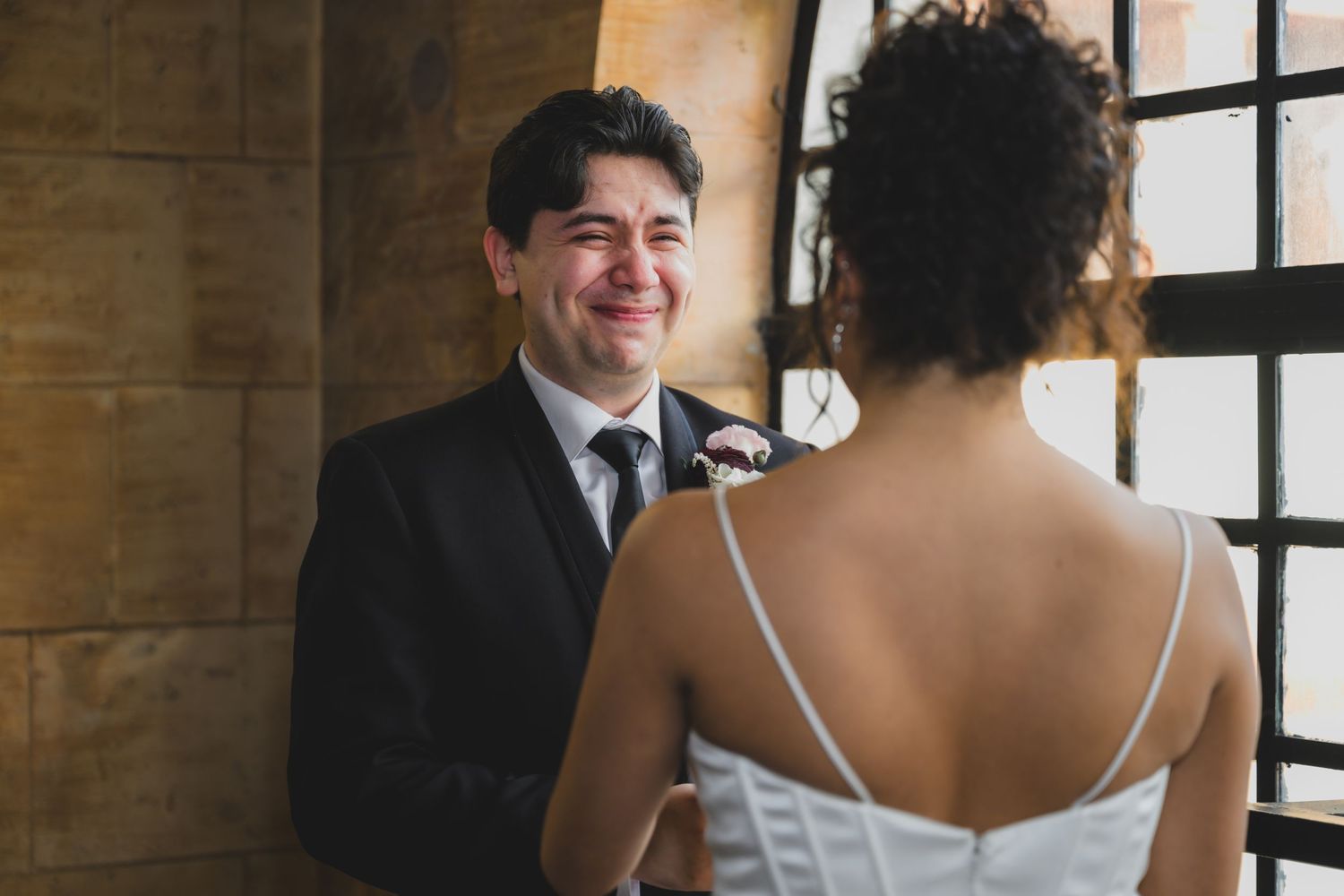 Groom smiling joyfully at bride in white dress during intimate wedding ceremony indoors.