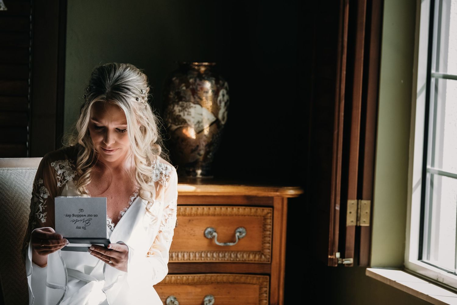 Blonde bride in white robe reads a heartfelt letter by window on wedding morning.