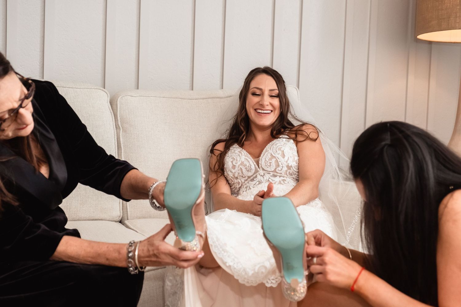 Bride laughing joyfully as bridesmaids help put on teal wedding heels while seated in white chair.