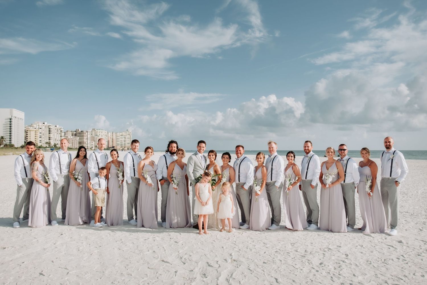 Large wedding party poses together on a white sandy beach under a blue sky.