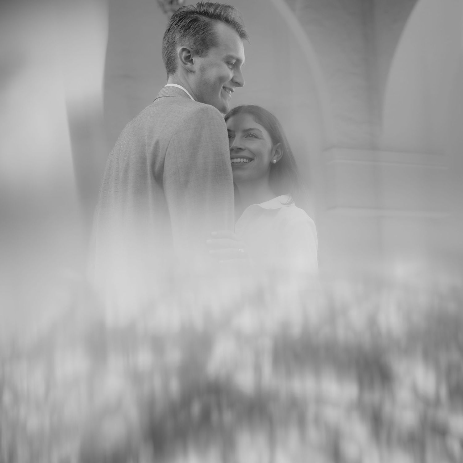 Black and white photo of a couple embracing and smiling among soft bokeh flowers.