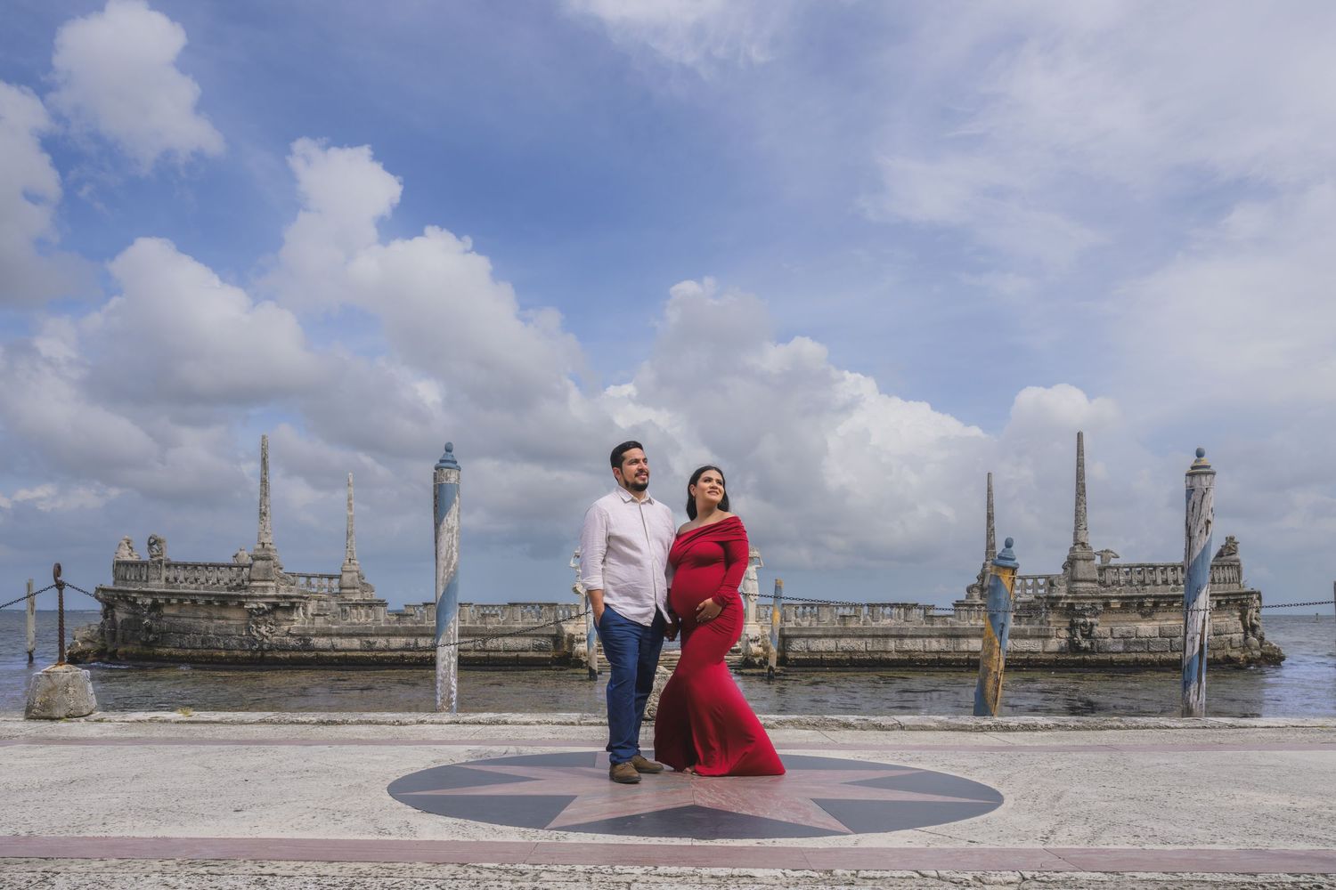 Couple posing at Vizcaya Museum waterfront terrace, woman in red maternity gown, man in white shirt, Miami.