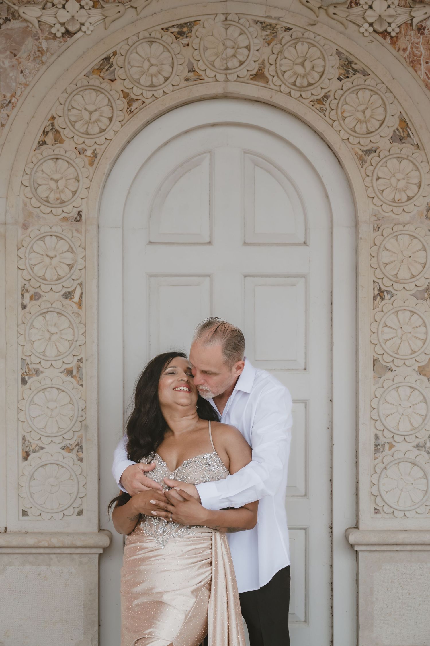 Couple embracing romantically in front of ornate arched door, woman in gold gown, man in white shirt.