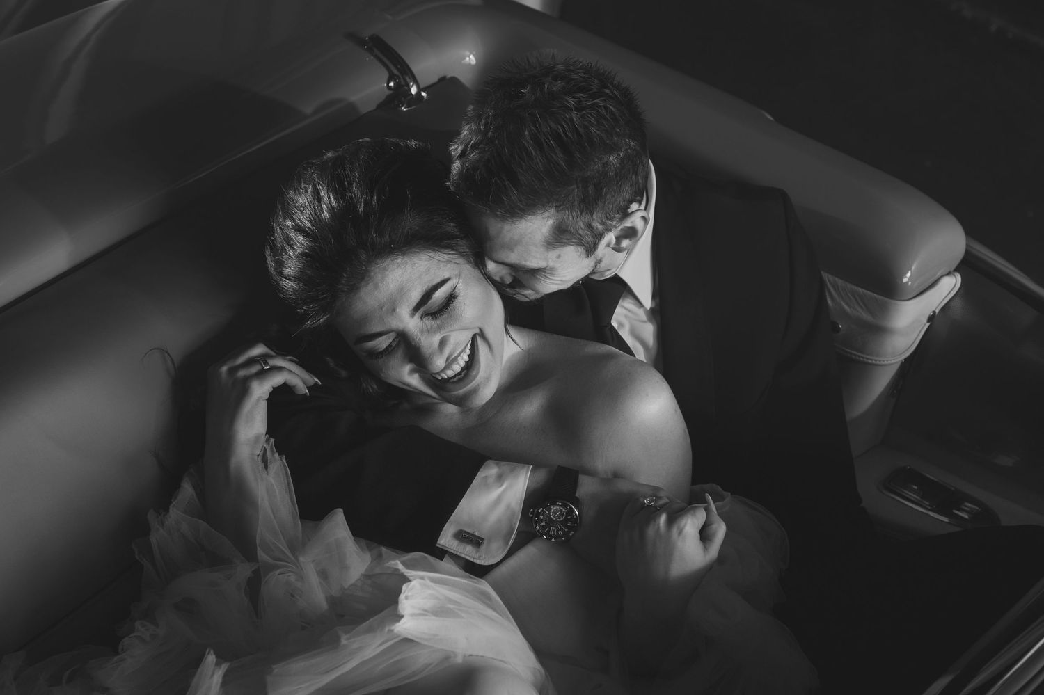 Black and white overhead photo of a laughing bride and groom sharing a romantic moment in a car.