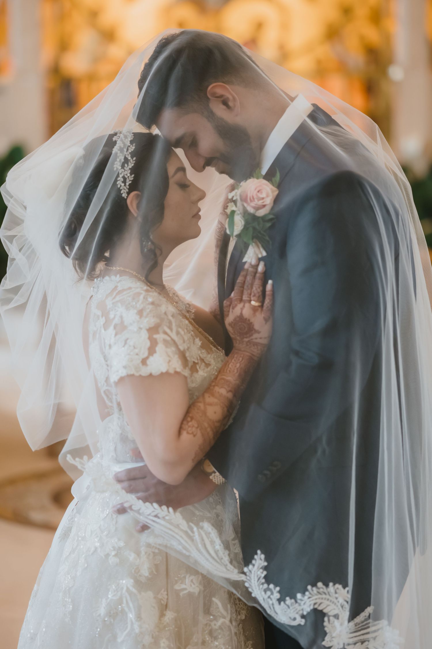 Bride and groom share an intimate moment under a lace wedding veil in a warmly lit venue.