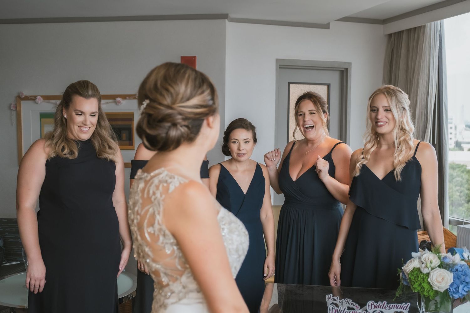 Bridesmaids in navy gowns react with joy seeing bride in lace wedding dress for first time.