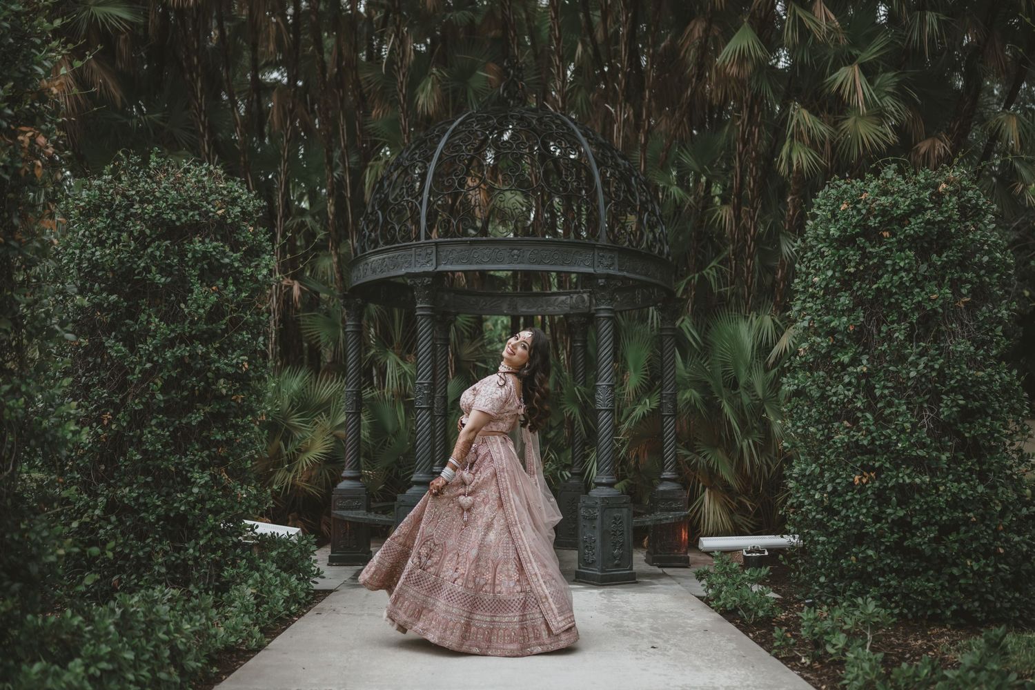 Indian bride twirling pink lehenga in lush garden gazebo surrounded by manicured hedges.