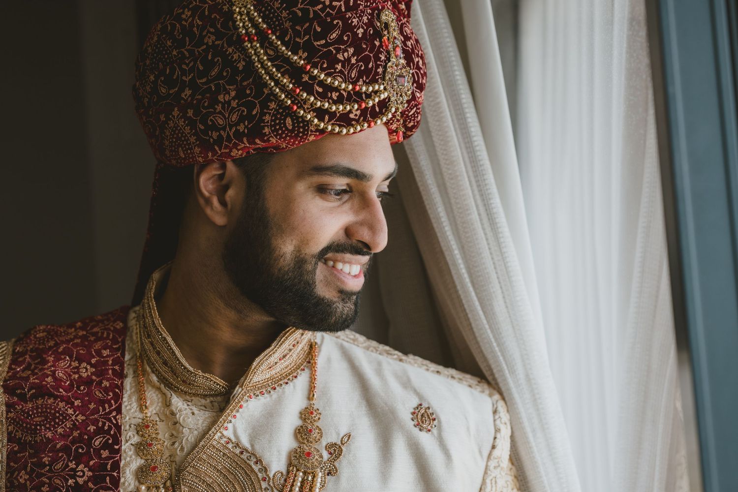 Smiling Indian groom wearing a red safa turban and embroidered sherwani on his wedding day.