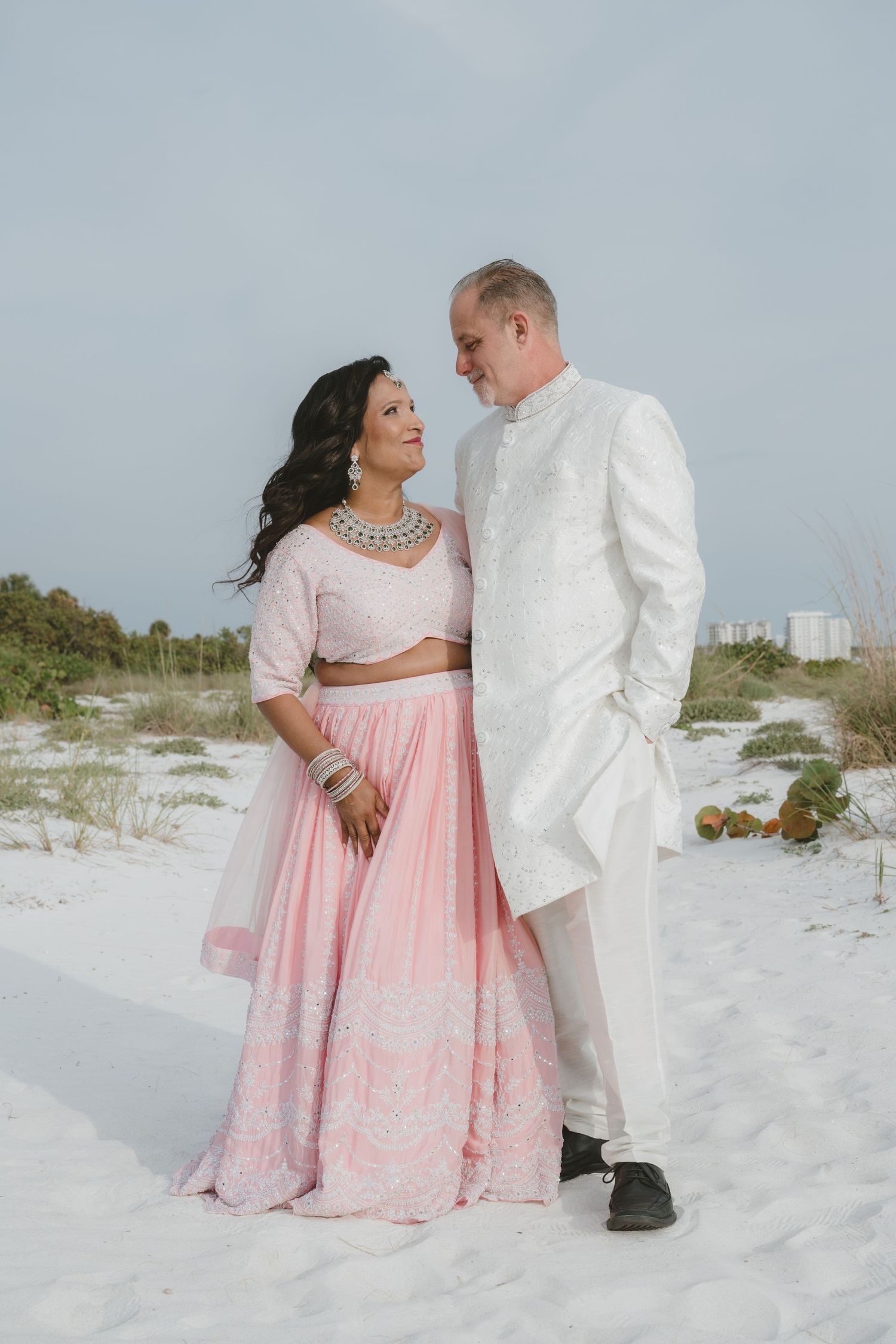 Couple in Indian wedding attire on white sand beach, woman in pink lehenga and man in white sherwani.