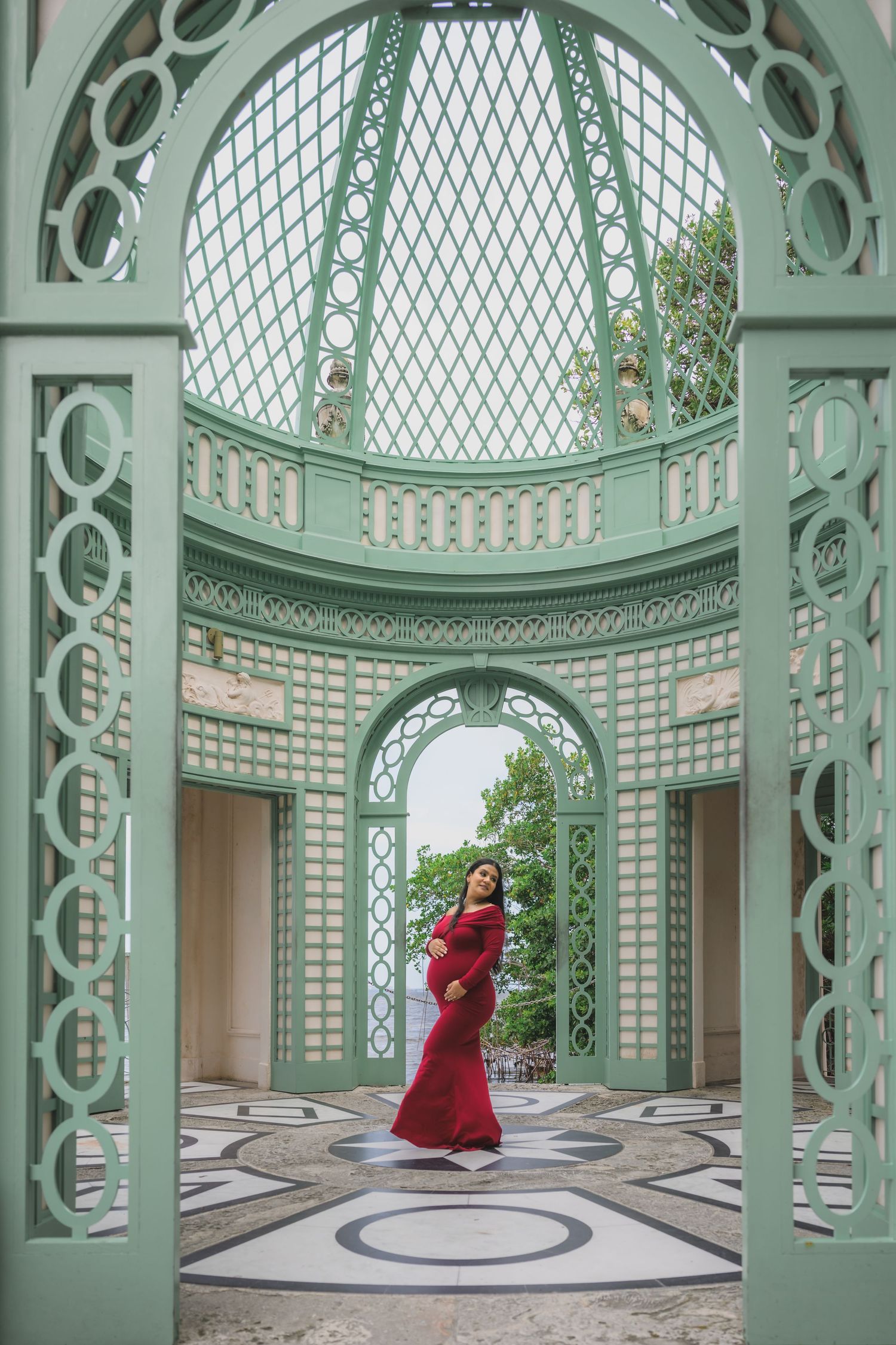 Pregnant woman in red gown poses inside ornate green lattice pavilion with arched doorways and domed ceiling.