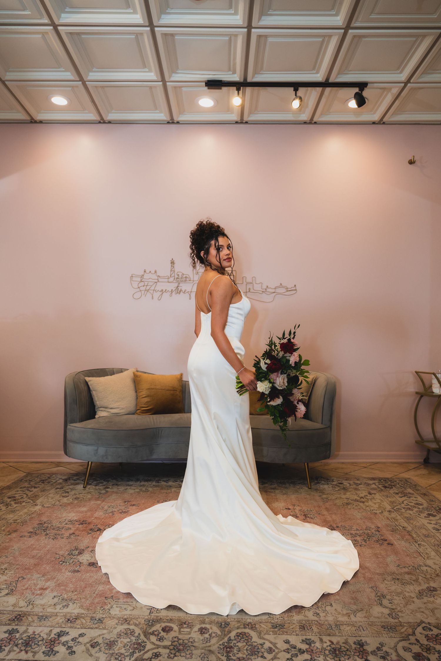 Bride in elegant white satin gown with train holds floral bouquet, posing in front of pink wall.